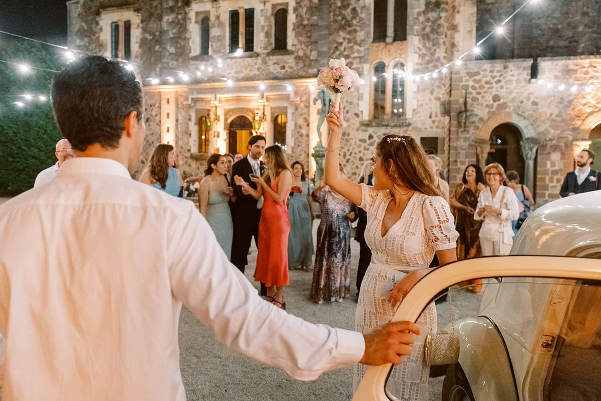 Bride raises bouquet at vintage car send-off as guests cheer beneath festoon lights at chateau courtyard