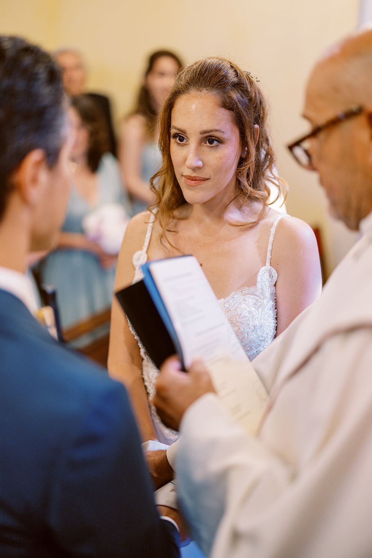 Bride in beaded spaghetti-strap gown watching officiant read from ceremony booklet during indoor ceremony