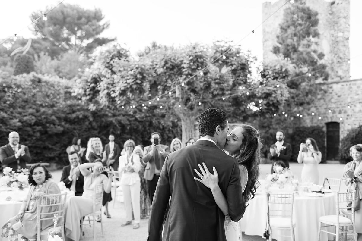A black-and-white image of a bride and groom sharing a kiss during what appears to be their first dance or a celebratory moment at an outdoor evening reception. The couple is photographed from behind and slightly to the side, with the bride's hand placed on the groom's back — she wears a lace-detailed gown and the groom is in a dark suit jacket. Approximately 15–20 guests surround the dance floor, many clapping, smiling, and photographing the moment on phones; chiavari chairs and round tables with floral centerpieces are visible on either side. The setting is an outdoor courtyard of a stone castle or historic fortified venue, with festoon bulb lights strung overhead adding warm tonal highlights against the dark foliage in the background. The image is a wide environmental shot with strong mid-tone contrast, capturing the energy of the crowd and the architectural stone tower in the upper right.