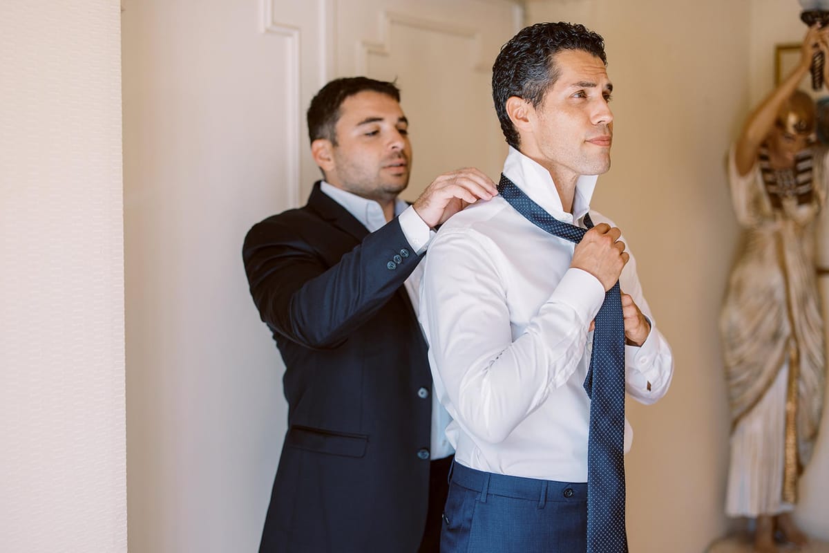A getting-ready moment captured indoors showing two men, with one helping the other adjust a navy blue polka-dot tie. The groom is wearing a white dress shirt and navy blue trousers, while the man assisting him wears a dark navy suit jacket over a light blue shirt. The setting appears to be inside a venue with cream-colored walls, and a decorative gold and cream figurine sculpture is partially visible in the background to the right. The shot is a medium portrait framing both men from approximately the waist up, with natural light illuminating the scene from the left.