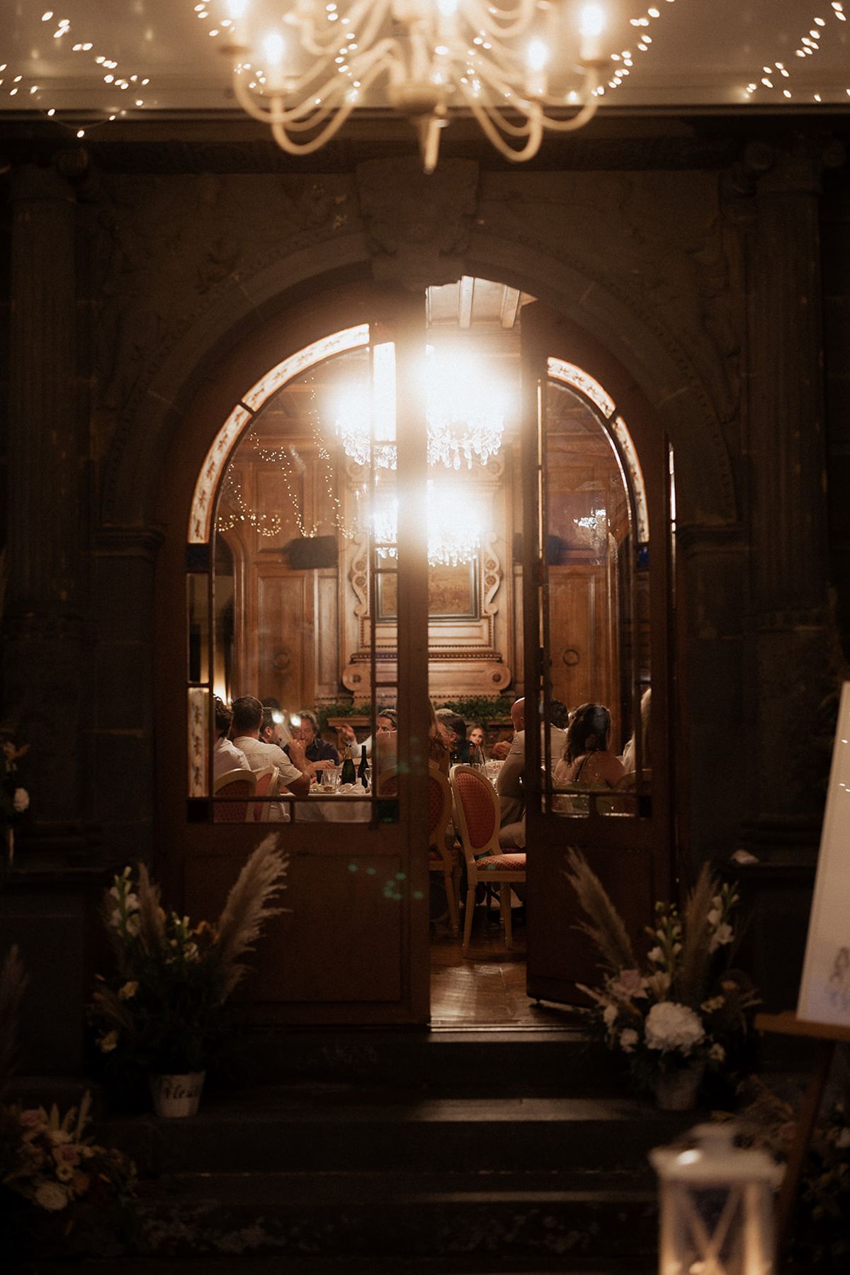 View through arched doors into fairy-lit reception dinner with pampas grass entrance arrangement