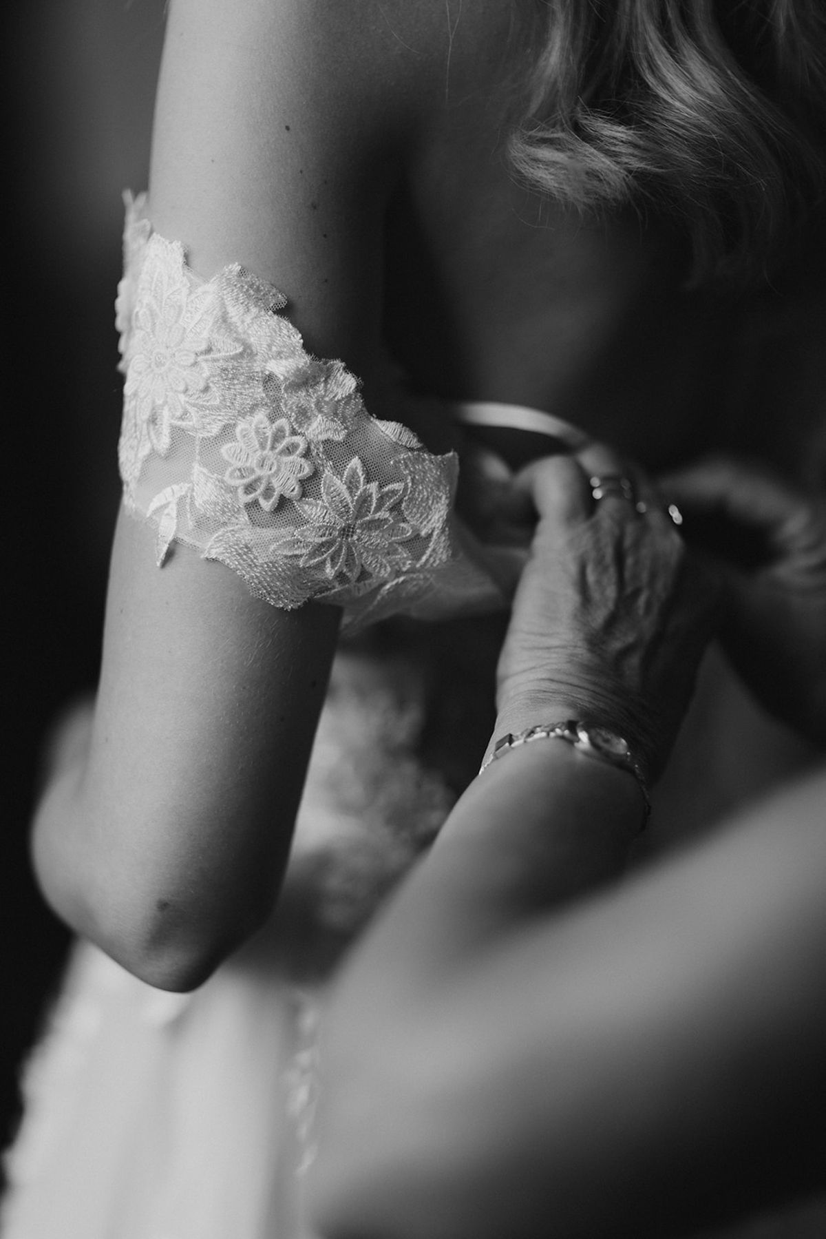 A black-and-white close-up detail shot captured during bridal preparations, showing an older person's hands — wearing a delicate bracelet and a ring — adjusting a lace garter on the bride's thigh. The garter features floral embroidery on a sheer tulle base. The bride has wavy blonde hair visible at the top of the frame, and the background is softly blurred with dark tones, drawing full focus to the garter and the hands. The high contrast and shallow depth of field emphasize the intergenerational moment of assistance.