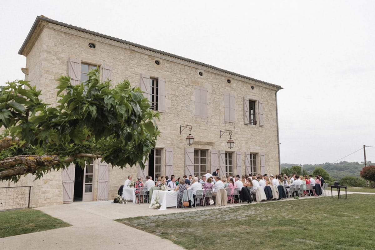 60 guests seated at long tables against Provencal stone farmhouse with taupe shutters