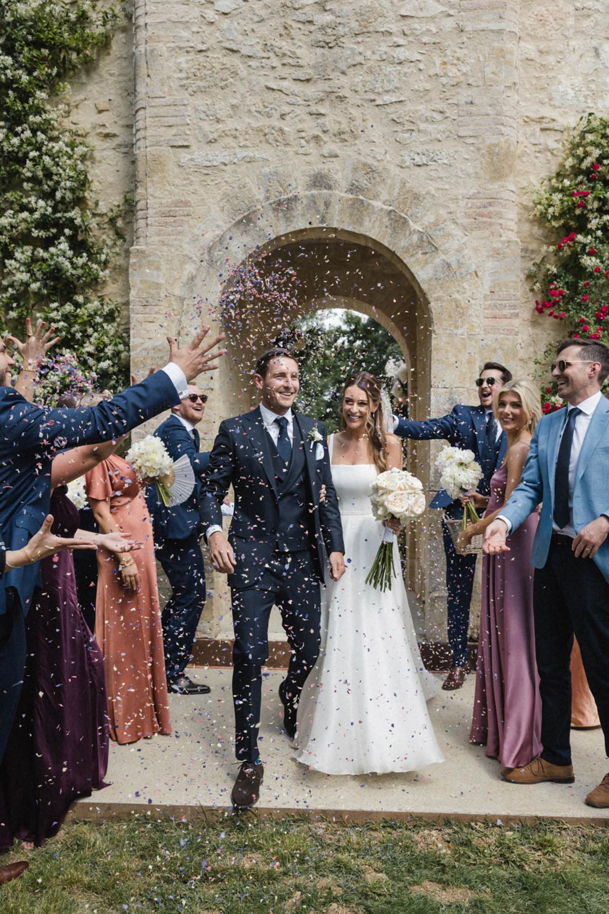 Bride and groom walk through confetti exit under stone archway with guests throwing colourful paper confetti