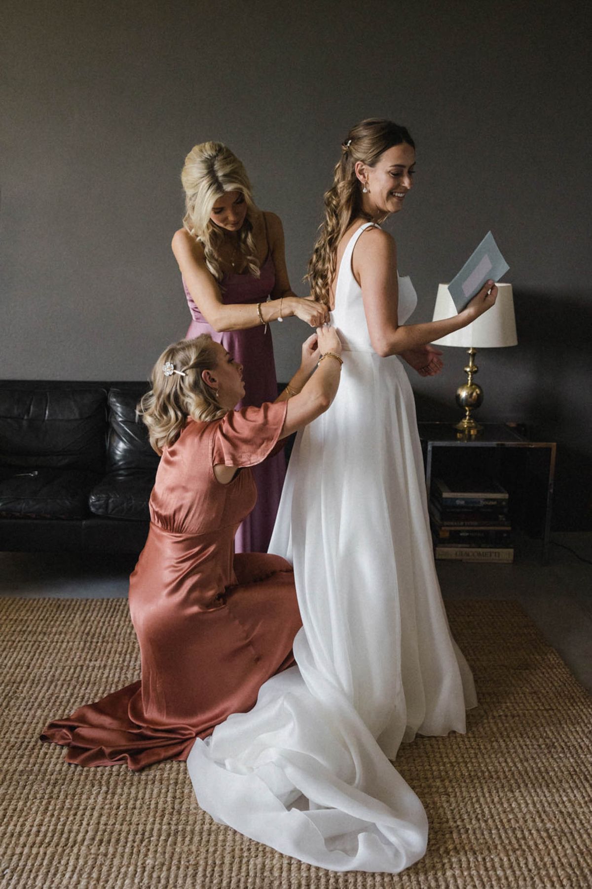 Bride smiling while two attendants in terracotta and mauve dresses fasten her gown in a darkly styled room