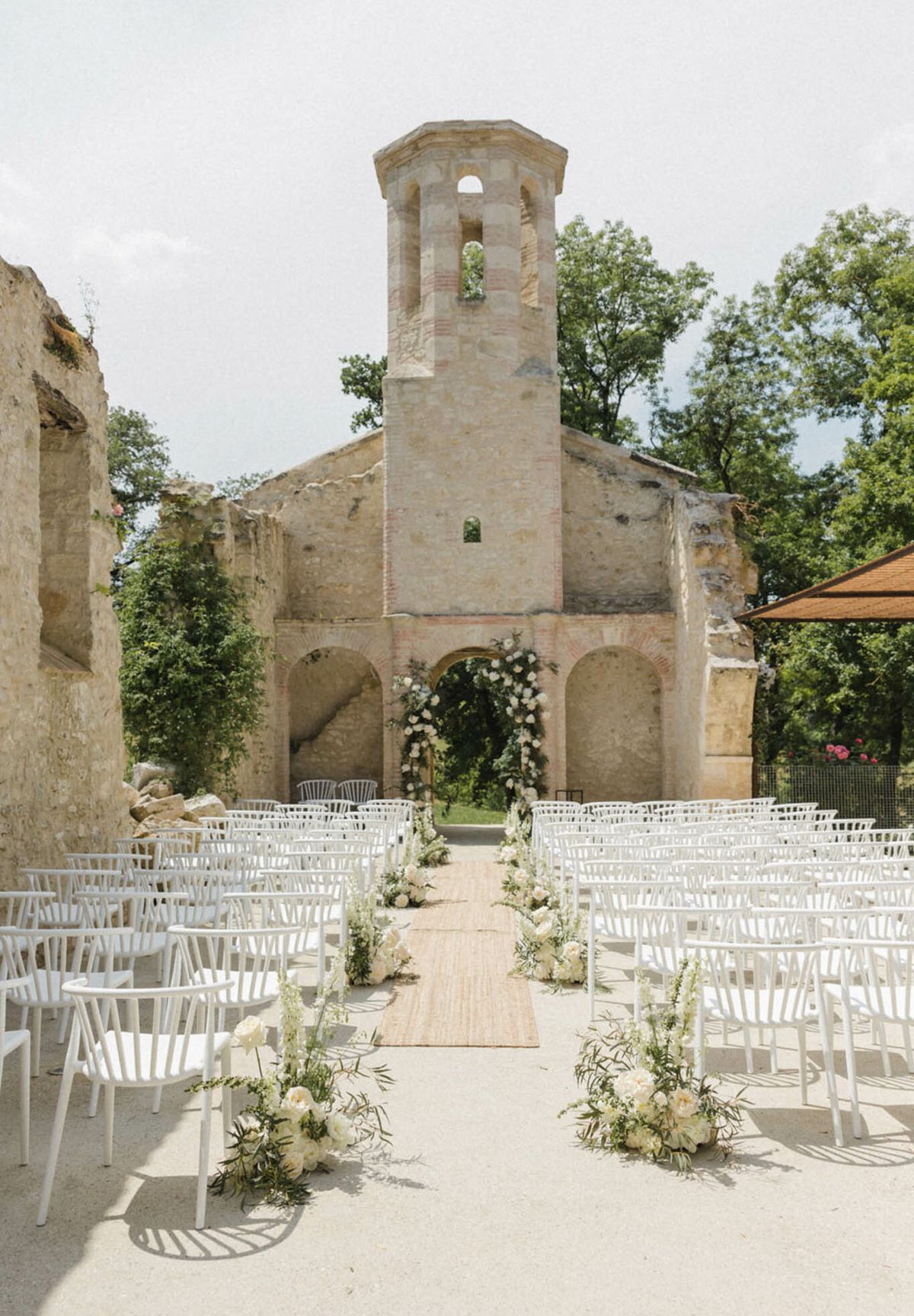 Ceremony setup in roofless stone chapel ruin with white floral arch, peony aisle clusters, and jute runner