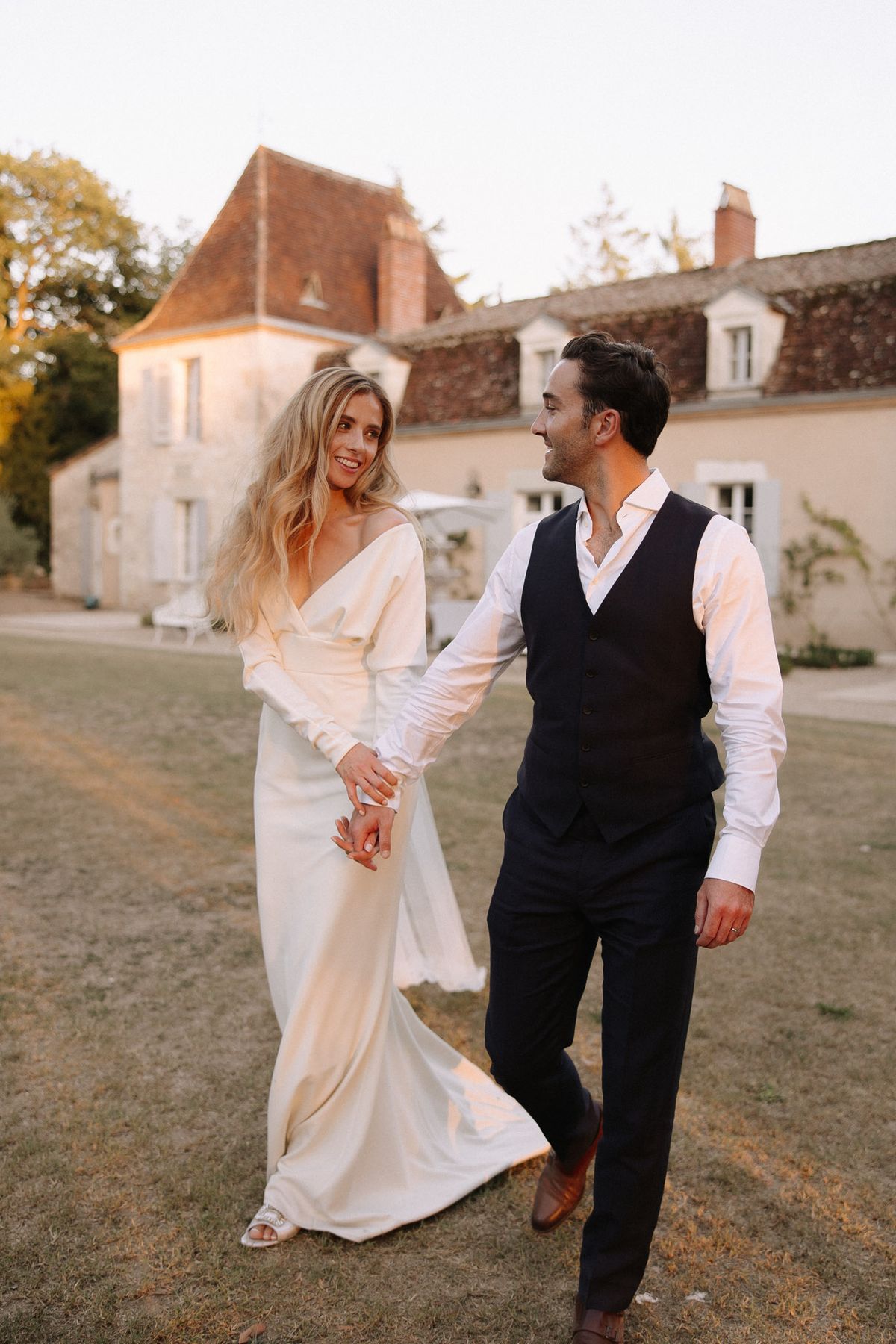 Couple walking hand in hand at golden hour bride in ivory satin slip gown groom in navy waistcoat at manor estate