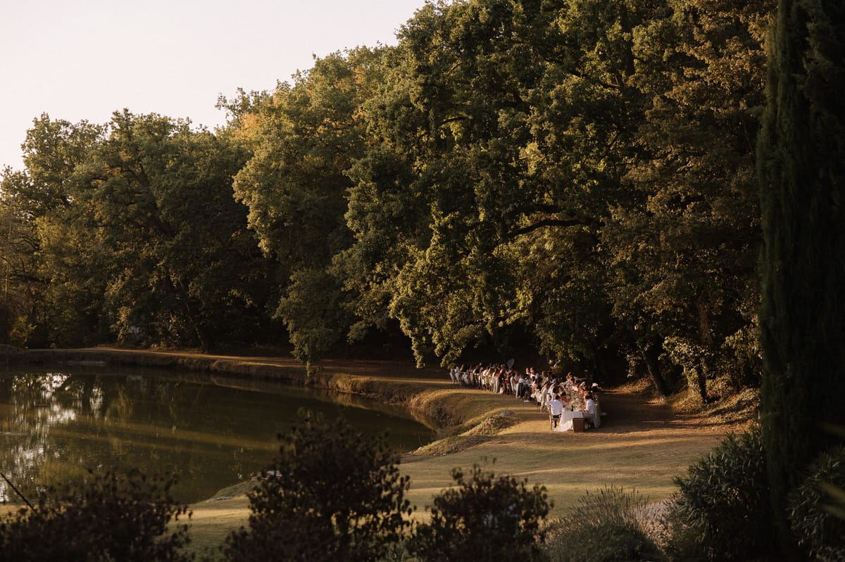 An outdoor wedding reception dinner is taking place along the bank of a calm ornamental lake, with approximately 40–60 guests seated at what appears to be a single long table running parallel to the water's edge. The setting is a large private estate or château grounds, with the table positioned on a flat lawn strip between the lake and a dense line of mature oak trees. Warm golden-hour light bathes the scene, casting long shadows across the grass. Table linens appear white or ivory, and floral centerpieces in warm, earthy tones are faintly visible at the near end of the table. The overall styling reads as classic and natural, making use of the landscape as the primary decorative element. This is a wide establishing shot taken from an elevated vantage point, showing the full length of the dining setup within its landscape context. Potential venue feature image.