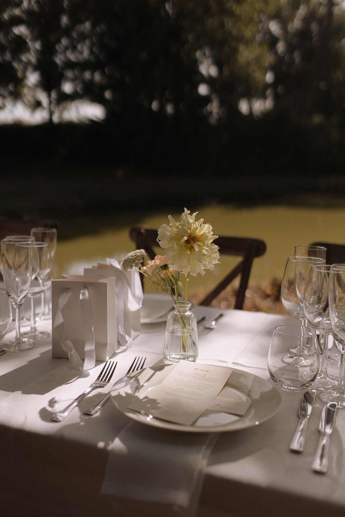 Reception table place setting with white plates, crystal glasses, cream dahlia bud vase, and printed menu card