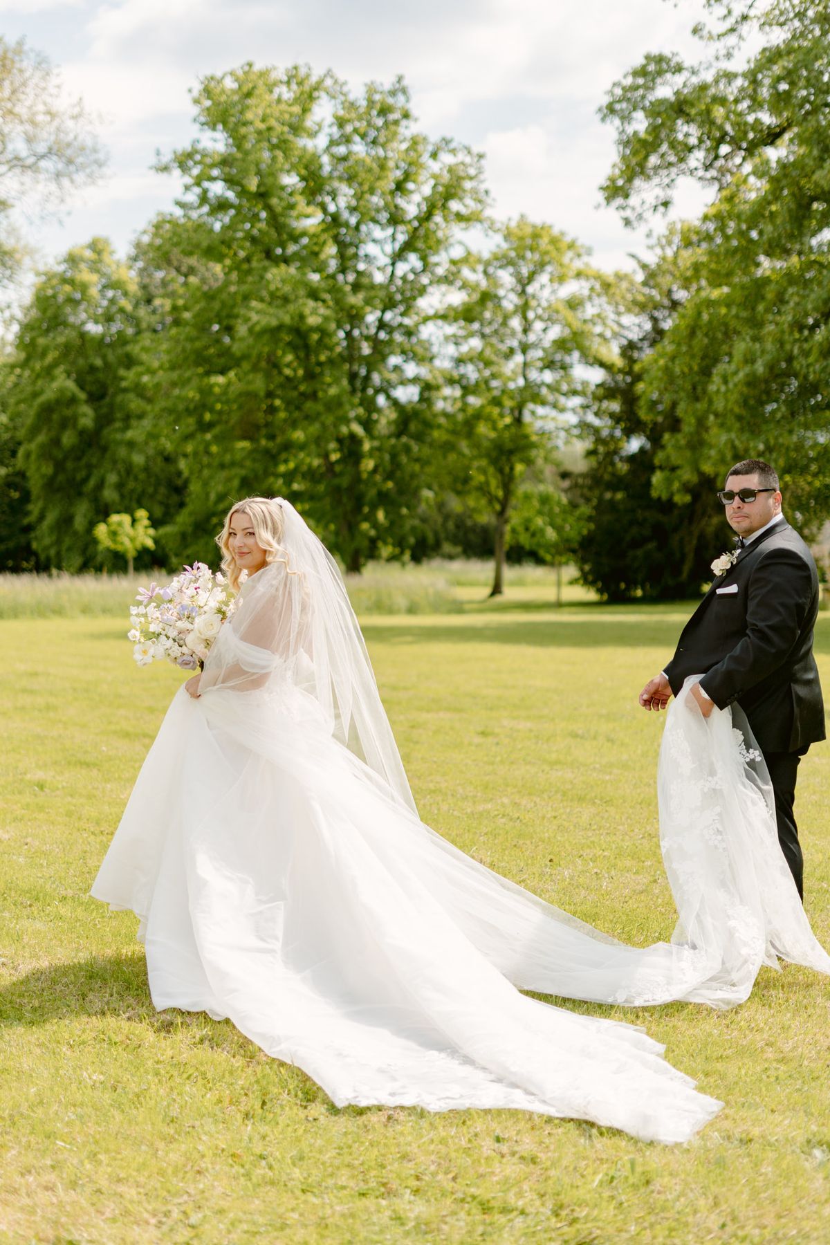 Groom holding bride's lace-edged train on lawn, bride with lavender and white bouquet glancing back