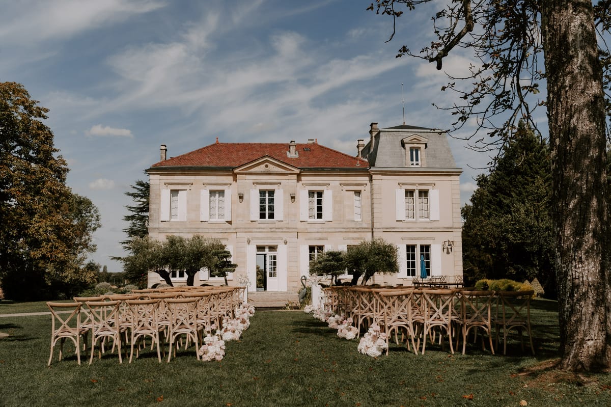 Crossback chairs facing limestone manor with blush and white floral aisle clusters beside olive trees