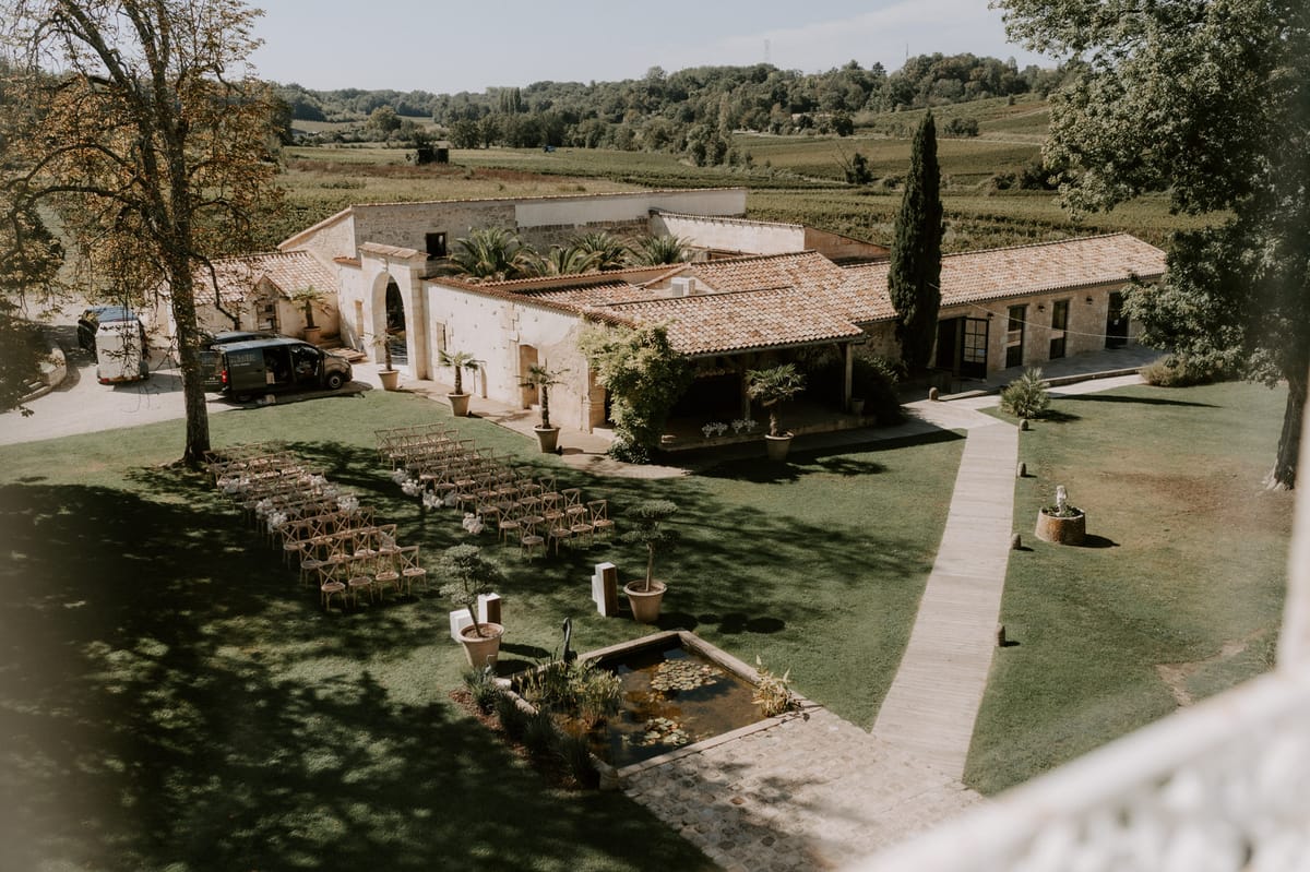 Elevated view of stone estate with outdoor ceremony chairs on lawn, lily pond, and vineyards in background