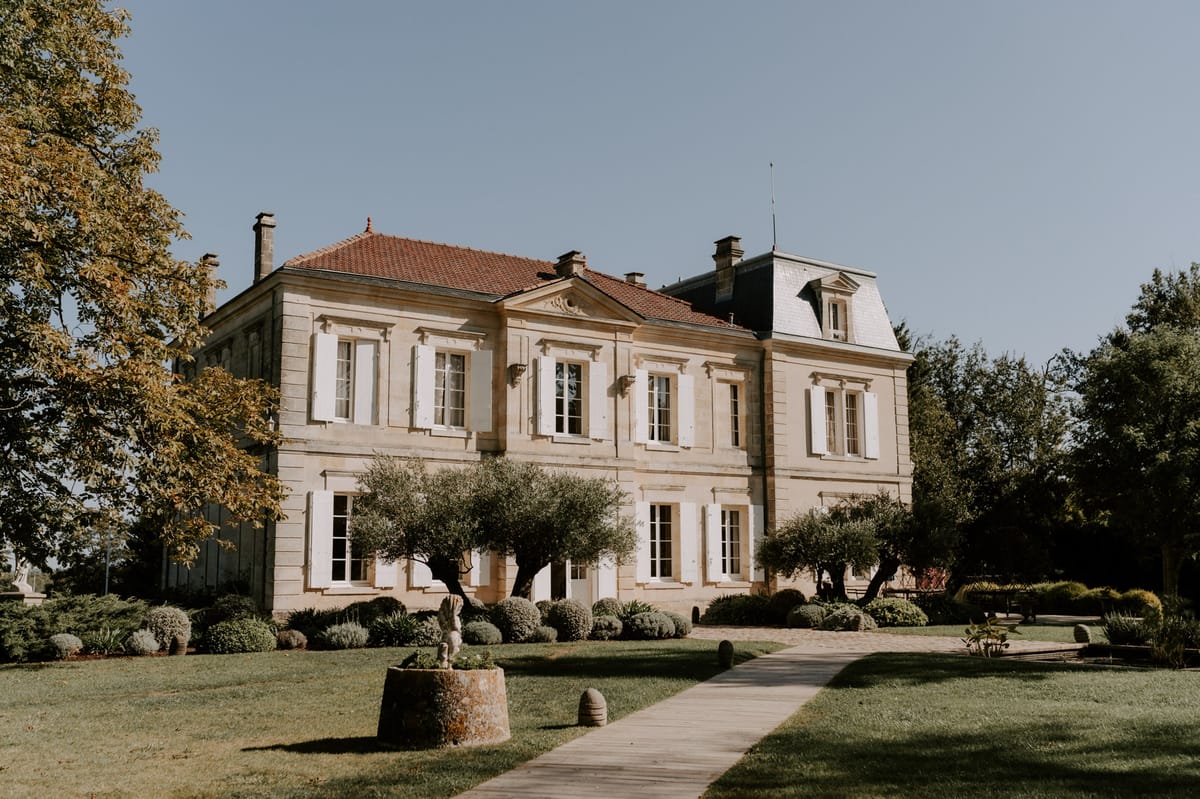 Golden limestone French chateau with terracotta roof, white shutters, trimmed olive trees and topiary along boardwalk path
