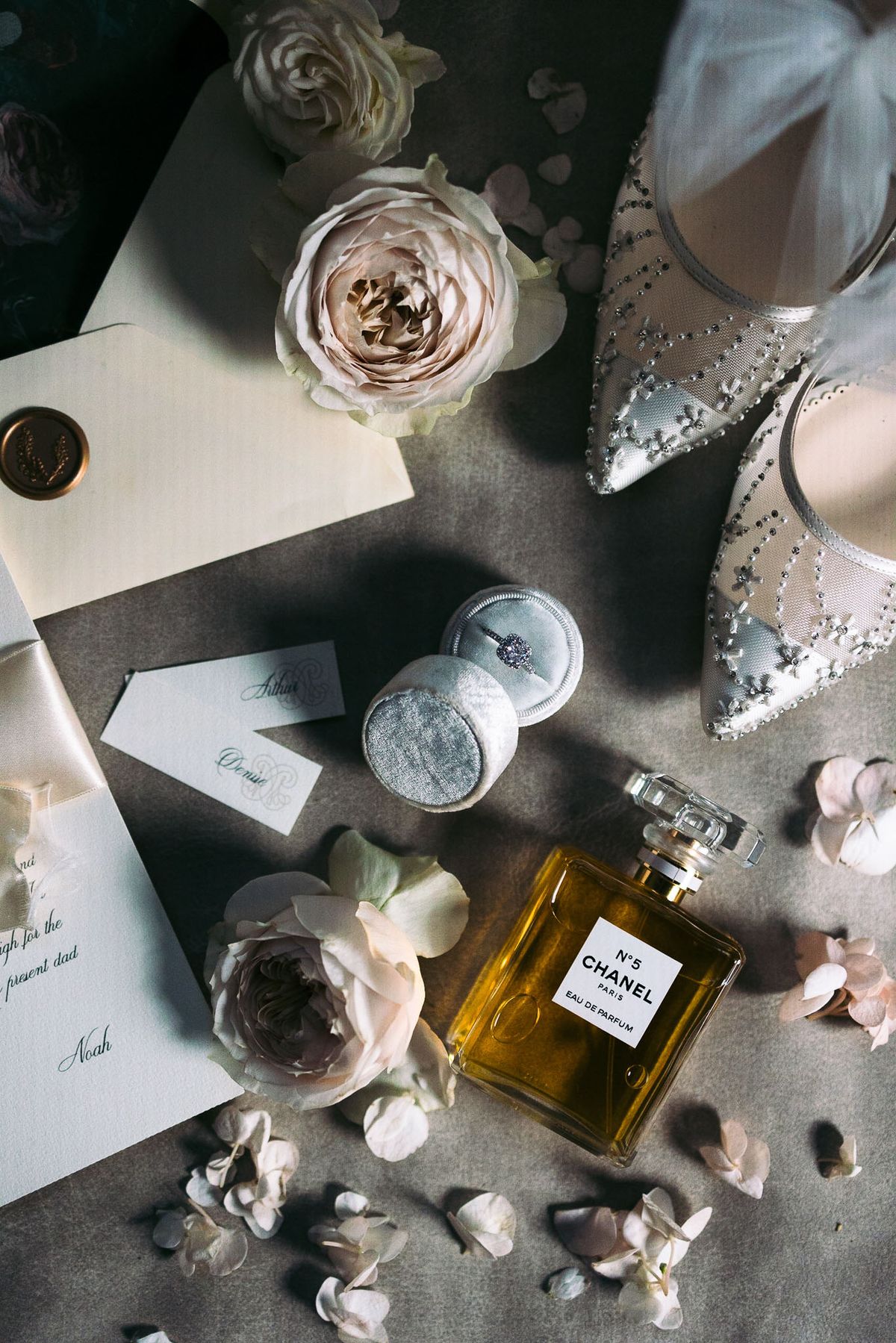 A flat lay detail shot of bridal accessories arranged on a grey surface. The items include a pair of white pointed-toe heels embellished with pearls and crystal beading, a diamond solitaire engagement ring displayed in an open dusty blue velvet ring box, a bottle of Chanel No. 5 Eau de Parfum, cream ivory envelopes sealed with a gold wax seal, calligraphy place cards reading 'Arthur' and 'Denise', a handwritten letter signed 'Noah', and scattered blush garden roses and loose white flower petals. The overall styling palette is grey, blush, ivory, and gold, with a classic and refined aesthetic.