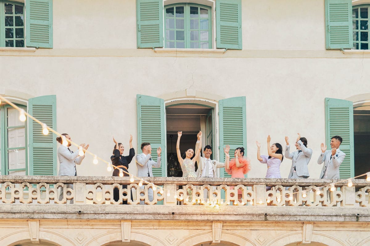 Bride and groom celebrate with guests on a chateau stone balcony with arms raised under string lights