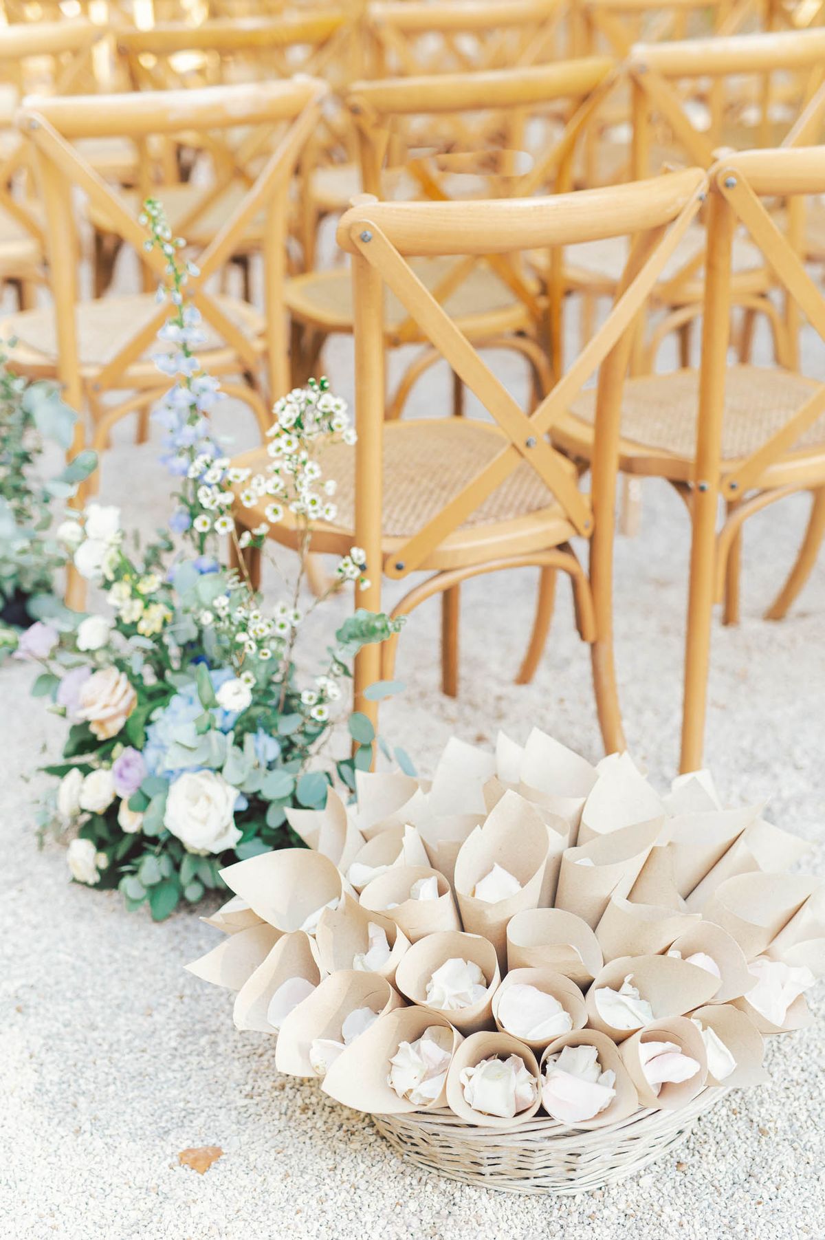 Wicker basket of kraft confetti cones beside blush rose, blue hydrangea, and delphinium aisle arrangement