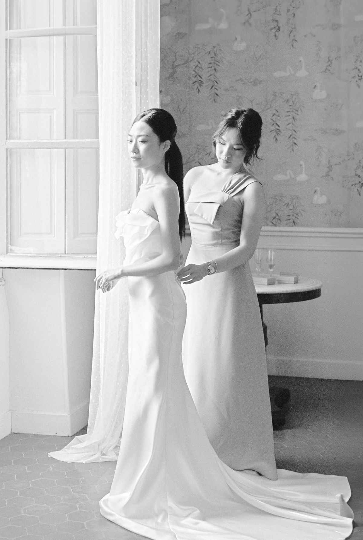 A black-and-white getting-ready portrait of two women in floor-length strapless gowns with structured bodice details and long trains, both wearing their dark hair up or pulled back. One woman stands facing away near a tall window with white painted shutters while the other adjusts the back of her dress, looking down. The room features decorative wallpaper with a hand-painted motif of swans and weeping willow branches, and a small round marble-topped side table in the background holding two champagne flutes and what appears to be a ring box. The image is shot in soft, high-key tones with gentle contrast, giving the scene a clean, modern aesthetic. The composition is a medium portrait shot emphasizing the interaction between the two women and the interior details of the room.