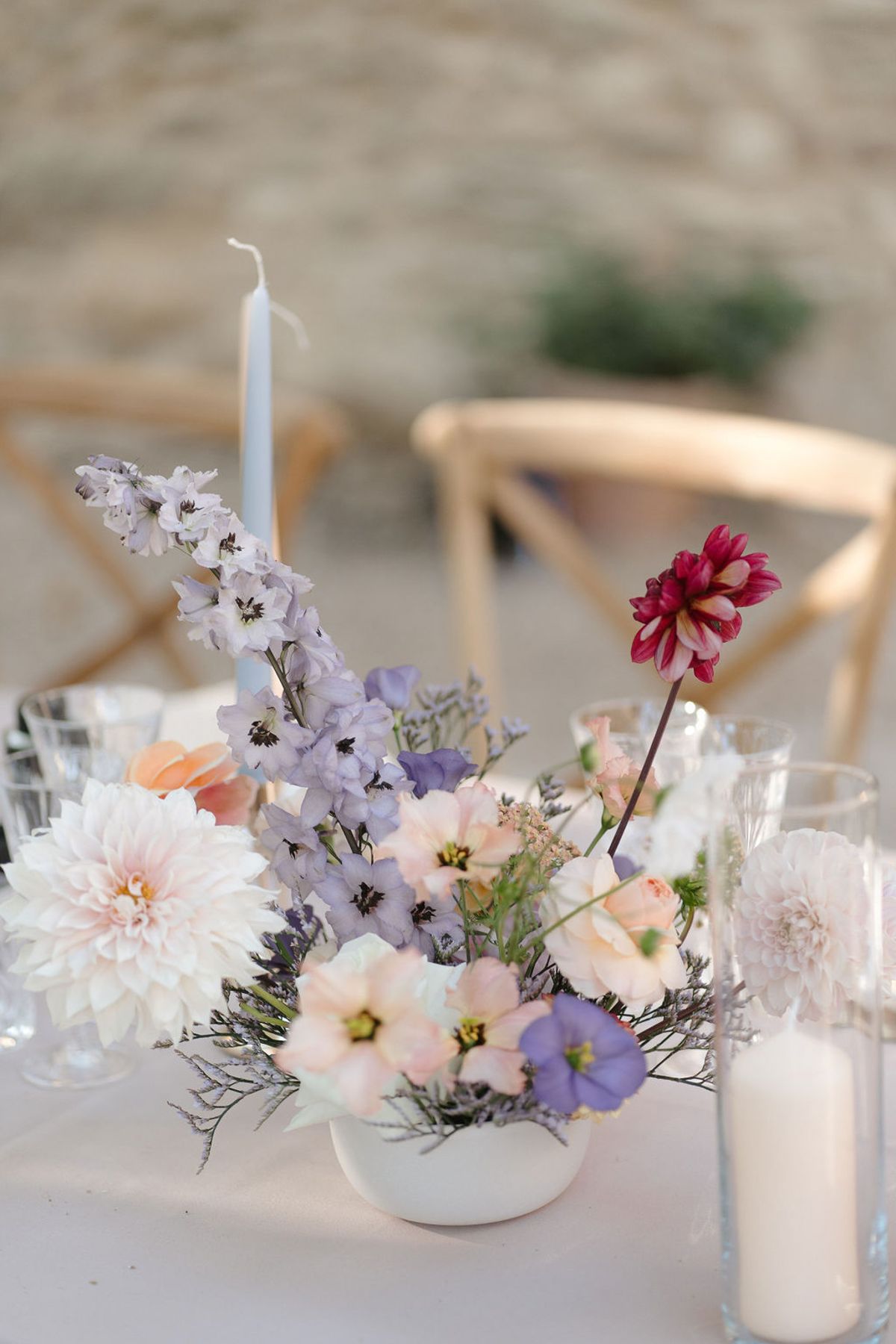 Garden-style centerpiece with blush dahlias, lavender delphinium, peach cosmos, and blue taper candle in white bowl