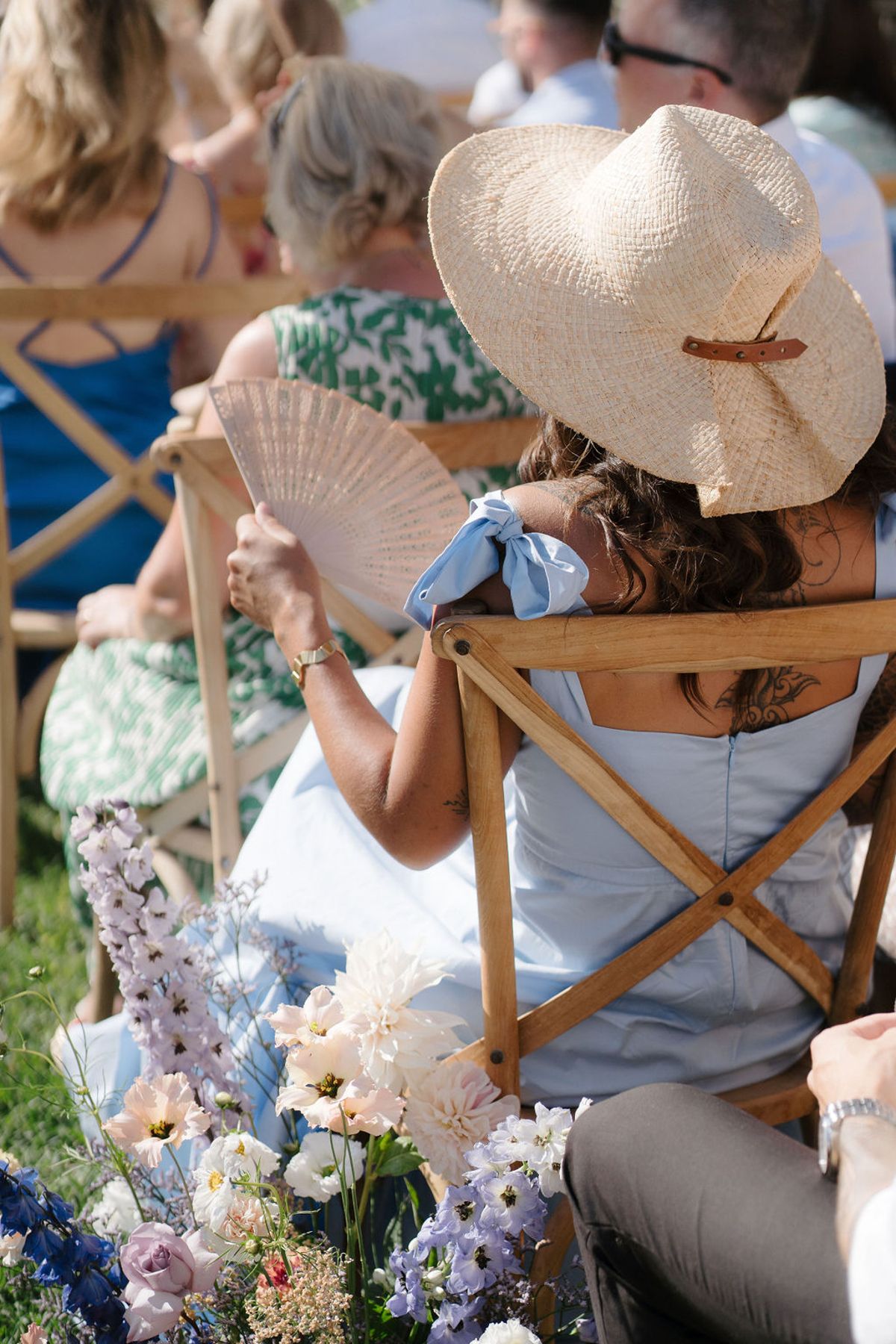 Wedding guest in pale blue dress and straw hat seated at outdoor ceremony with pastel floral aisle arrangements