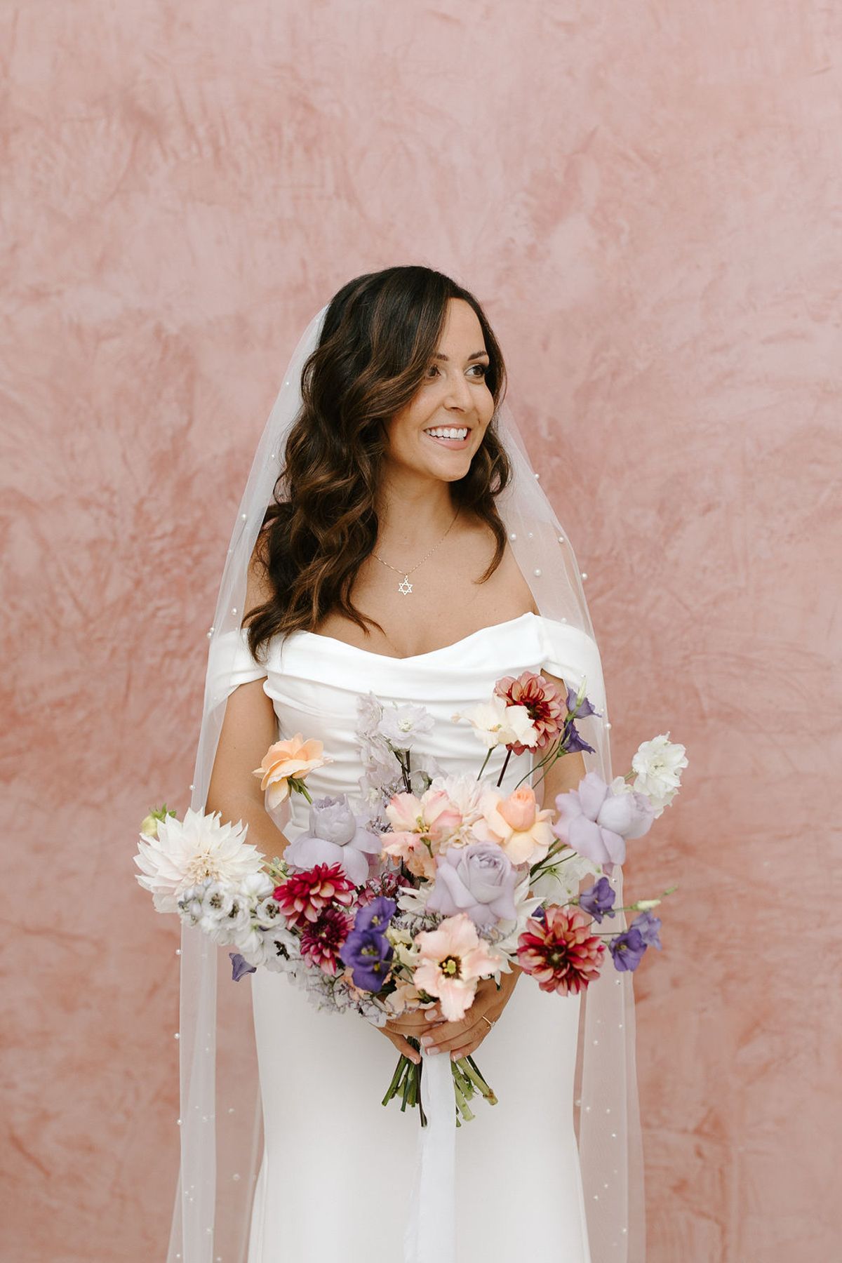 A bridal portrait of a single bride standing against a terracotta-pink textured wall. She wears a white off-the-shoulder satin gown with a draped neckline and a pearl-edged veil, and accessorizes with a Star of David pendant necklace. She holds a loosely arranged bouquet featuring lavender roses, peach roses, burgundy and white dahlias, purple lisianthus, and pink anemones. Her dark brown hair falls in loose waves over her shoulders. The shot is a mid-length portrait with the bride glancing slightly to one side and smiling.