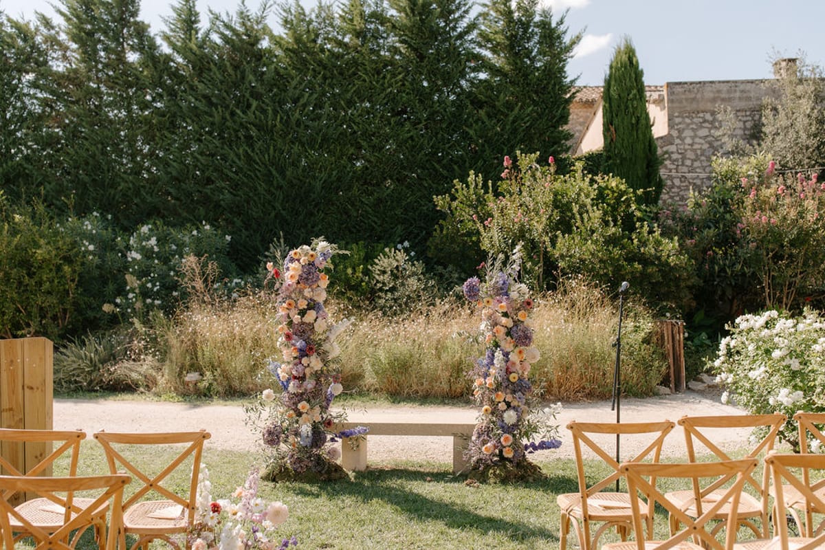 Outdoor garden ceremony setup with two tall floral column installations in purple, peach, and blush beside wooden cross-ba...