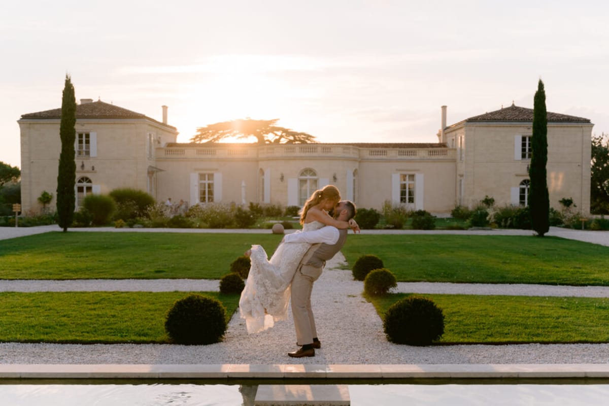 A couple portrait taken outdoors at golden hour, with the groom lifting and dipping the bride as they kiss on a gravel pathway in front of a French château. The bride wears a fitted lace white gown with a train, and the groom is dressed in light beige trousers and a white shirt. The formal garden features clipped box ball topiaries lining the gravel path and a reflecting pool in the foreground. The château is a classic two-storey limestone French country manor with symmetrical wings, white shuttered windows, and terracotta roof tiles, flanked by tall cypress trees. The sun sets directly behind the building, creating a warm backlit glow across the entire scene. A few guests are faintly visible near the château entrance in the background. Wide portrait shot. Potential venue feature image.