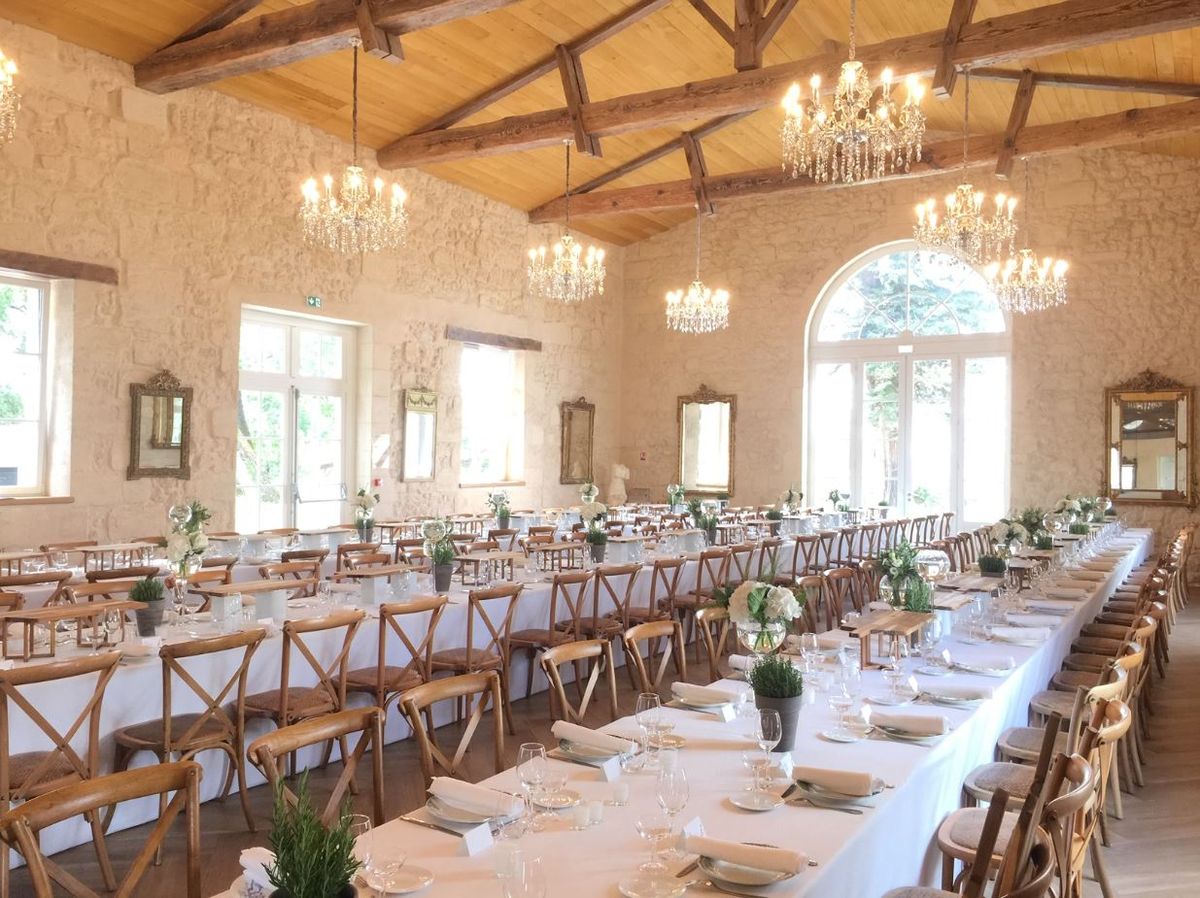 A wide shot of an indoor wedding reception room set for a large dinner, with no guests present. The room features exposed wooden beam vaulted ceilings, cream-painted stone walls, and multiple crystal chandeliers hanging at varying heights. Four gilt-framed mirrors are mounted along the walls between tall arched and rectangular windows. Long rectangular tables are covered in white linen and arranged in parallel rows, each set with white plates, folded white napkins, and multiple wine glasses. Centerpieces consist of small potted green herbs and low arrangements of white flowers, including what appear to be white roses and white hydrangeas, in glass vases. Seating is provided by natural wood cross-back chairs with woven seats. The decor palette is white, green, and natural wood, giving the room a rustic-classic feel. Potential venue feature image.