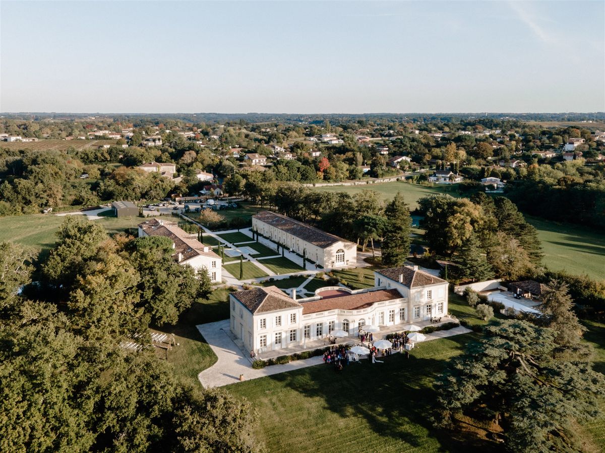 Aerial view of cream limestone chateau with formal garden, cypress trees, and guests on forecourt terrace