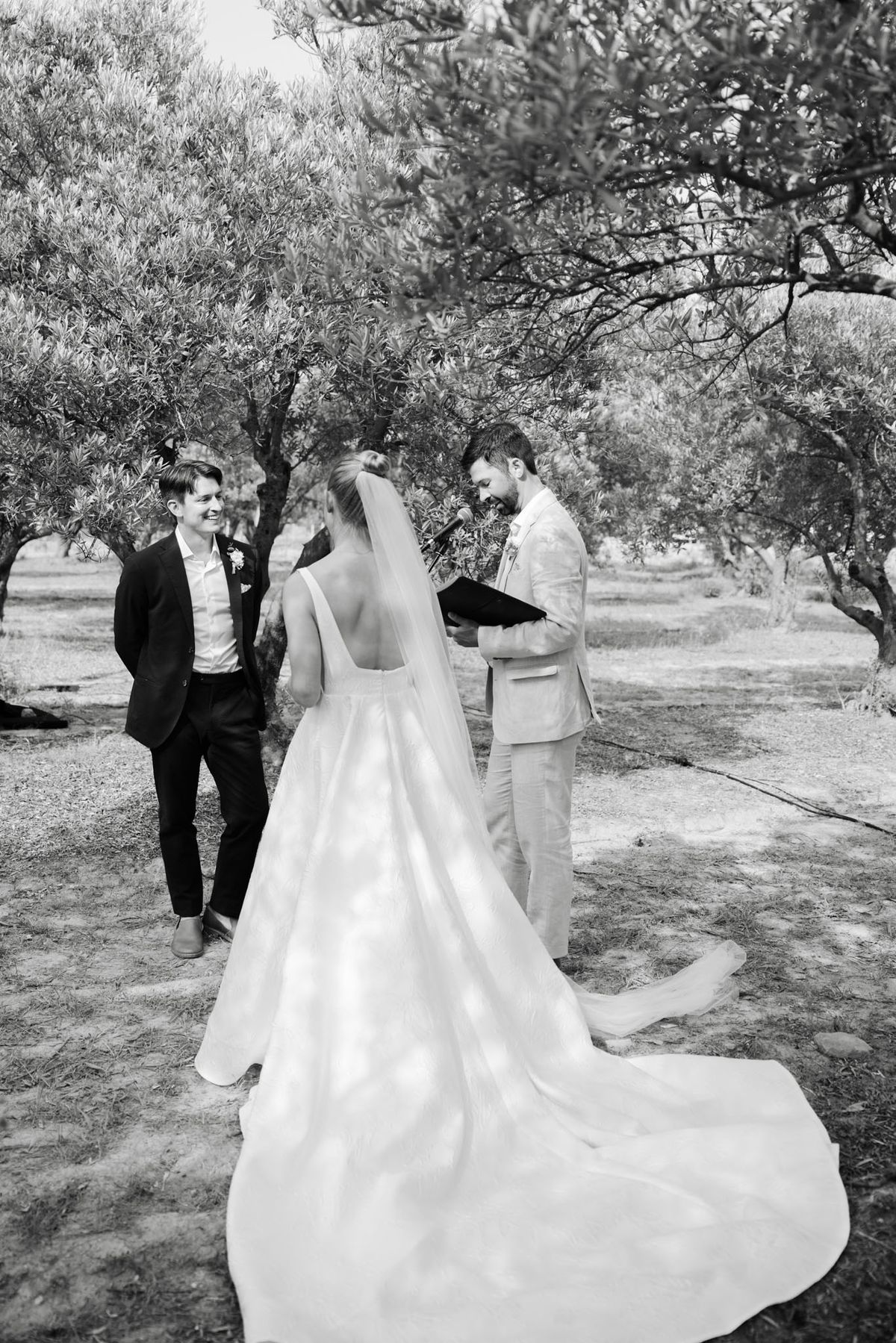 This black-and-white image captures an outdoor wedding ceremony taking place among a grove of olive trees. The bride is seen from behind, wearing a square-neck ball gown with a long cathedral train and a single-tier veil, her hair styled in an updo; she faces the groom, who wears a light-toned suit and holds an open ceremony booklet while speaking into a microphone. A second figure — likely the officiant or a groomsman — stands to the left in a dark suit with a boutonnière, smiling warmly toward the couple. The image is shot in high contrast black and white with bright highlights on the bride's dress and soft mid-tones throughout, and is framed as a medium wide shot taken slightly from above and behind the bride.