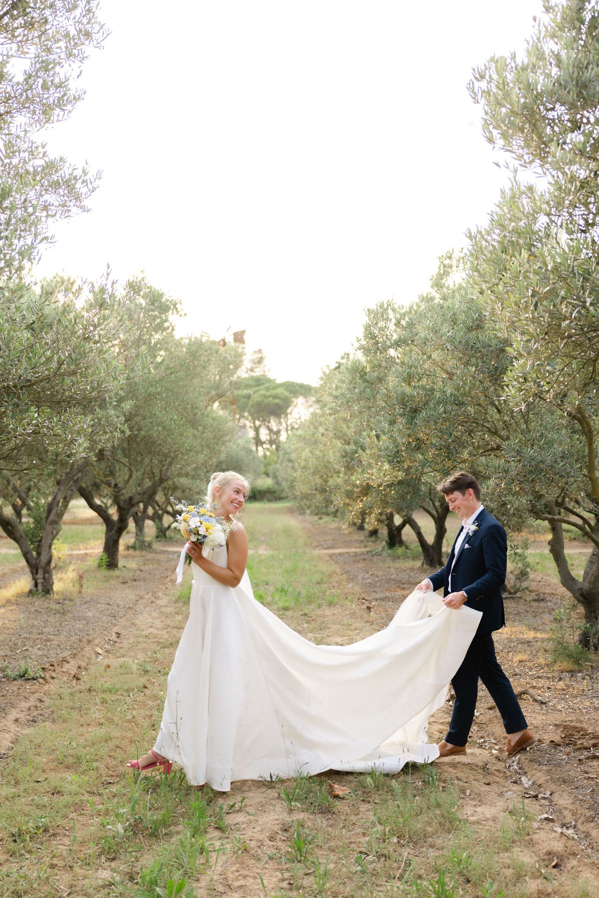 Groom carries bride's train as they walk through an olive grove, bride holding wildflower bouquet