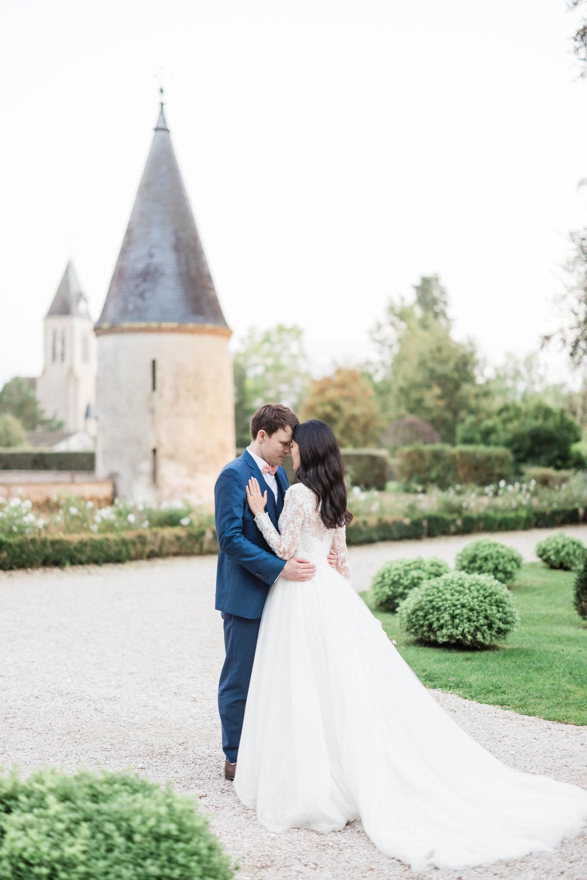 A couple portrait taken outdoors in the formal gardens of a French château, with a round stone tower featuring a conical slate roof visible in the soft-focus background. The bride wears a long-sleeve lace-bodice gown with a full ivory tulle skirt and an extended train, while the groom is dressed in a navy blue suit with a blush pink tie. The two are standing close together on a gravel path, foreheads nearly touching, with the groom's arm around the bride's waist. The garden features clipped round boxwood topiaries, neatly trimmed hedgerows, and beds of white flowering plants arranged in a classic French formal style. The composition is a mid-length portrait shot with the couple sharply in focus against the softly blurred château grounds.