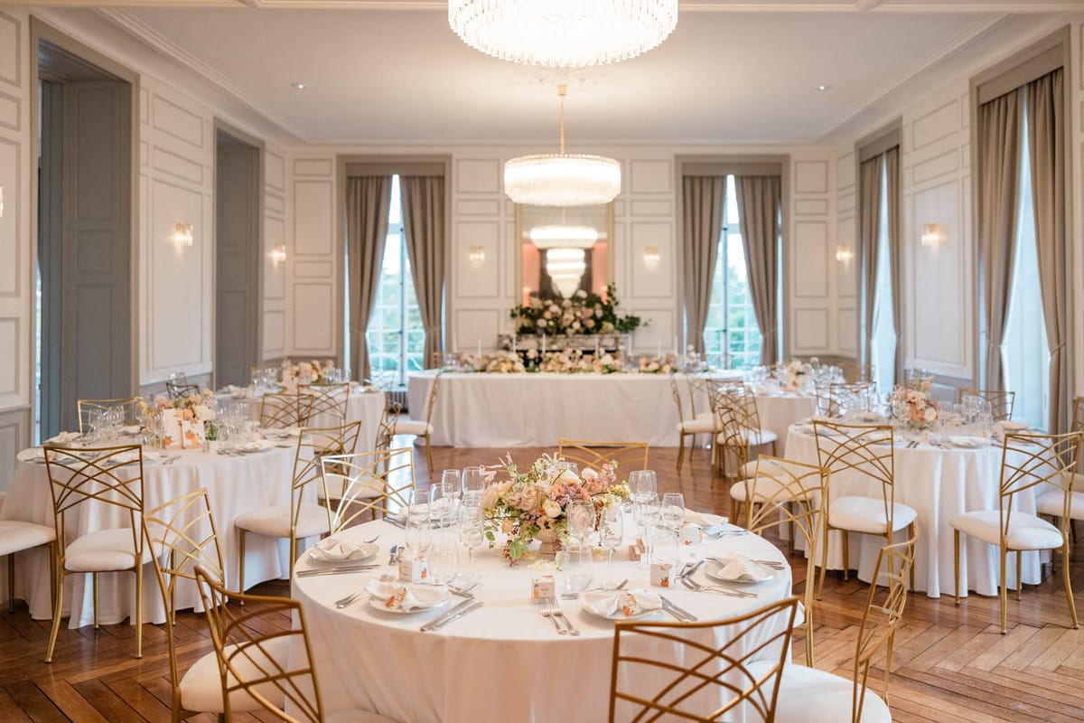 A wedding reception room setup shot inside a formal ballroom-style venue with white panelled walls, parquet flooring, and floor-length taupe curtains. The room features multiple round guest tables dressed in white floor-length linens, surrounded by gold geometric-frame chairs with ivory cushioned seats, and a long rectangular head table at the back of the room set against tall windows. Table centerpieces consist of low, lush arrangements of blush, dusty rose, cream, and mauve blooms including what appear to be garden roses, ranunculus, and trailing greenery, placed in natural wood-toned vessels. The head table features a more elaborate elevated floral arrangement with greenery and blush tones at its center. Two large crystal tiered chandeliers hang from the ceiling, supplemented by wall sconces that cast warm light throughout the space. Each place setting includes white folded napkins, silver cutlery, multiple wine glasses, and small decorative place cards. The overall decor palette is white, gold, and blush, with a classic French interior style. Wide-angle room shot with no people present. Potential venue feature image.