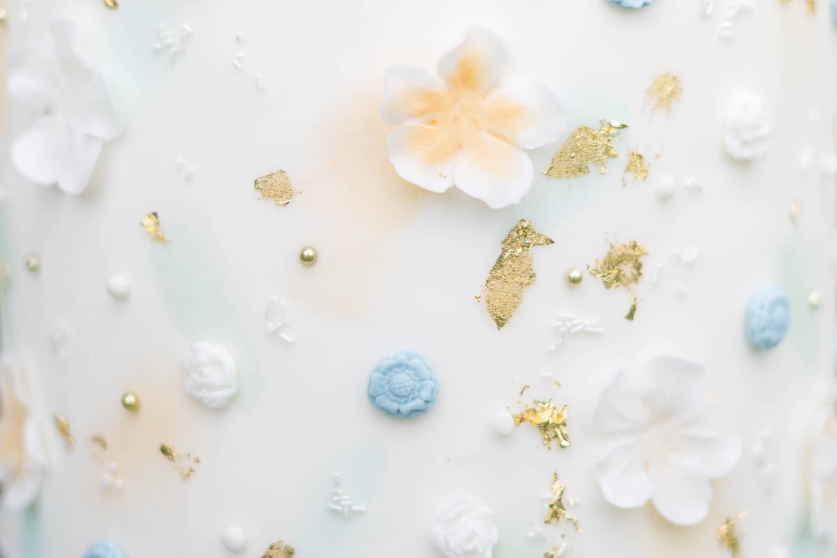 Close-up cake tier with sugar flowers, blue medallions, gold leaf, and pale watercolour brushwork