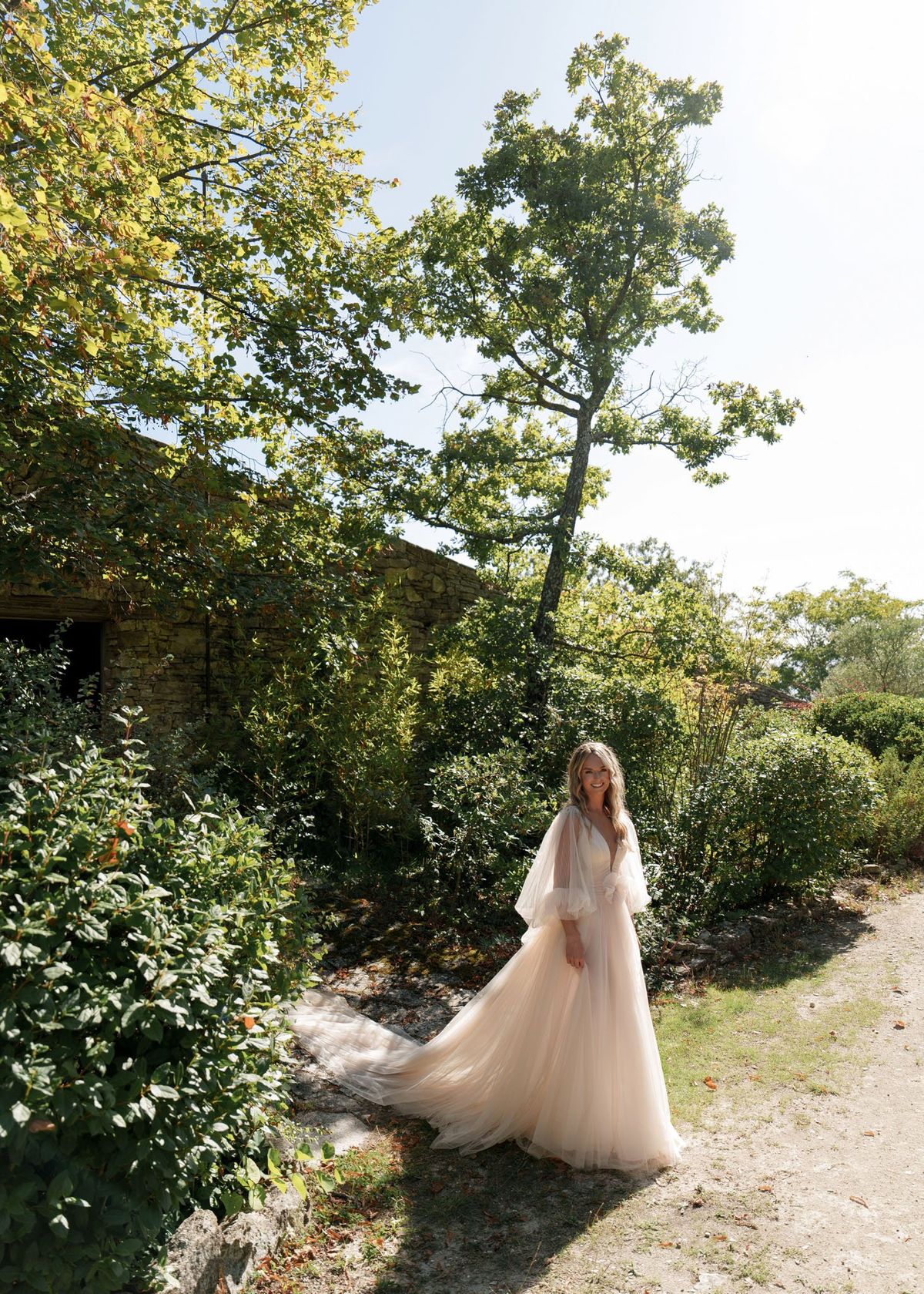 Bride standing on gravel path in blush tulle ball gown with flutter sleeves and long train beside stone building
