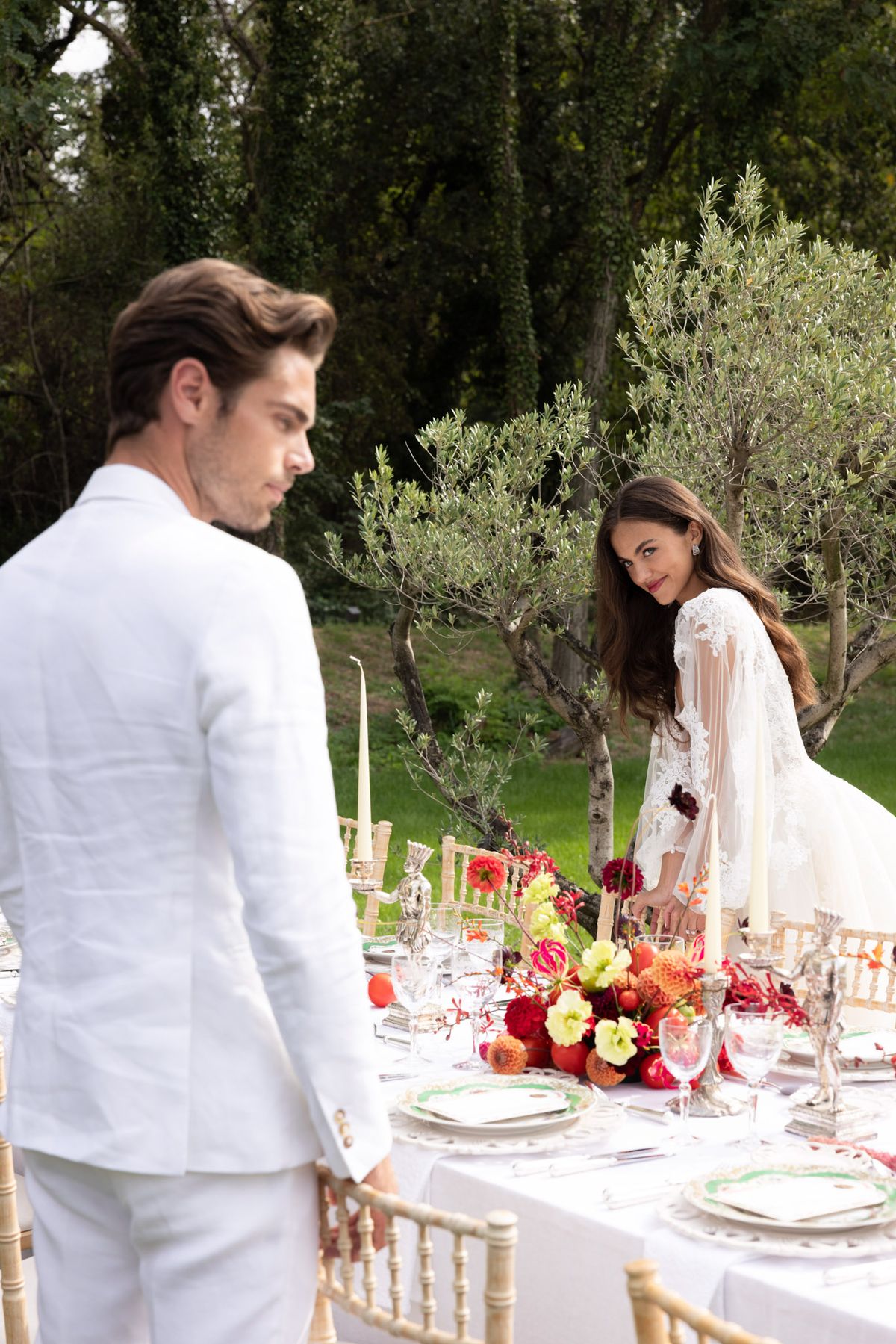 Bride in white lace gown and groom in white linen suit at garden reception table with crimson and orange dahlia centerpiece