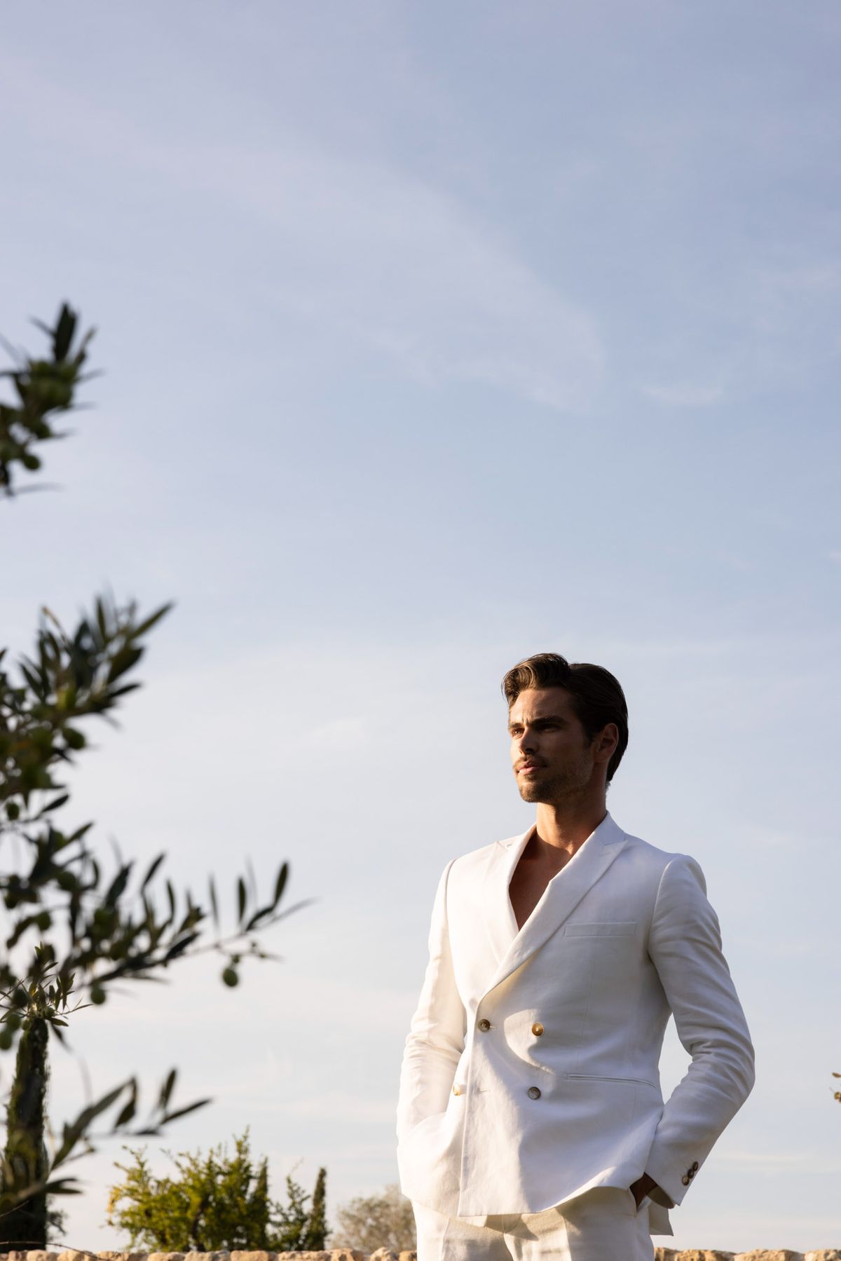 Groom in white double-breasted linen suit with gold buttons photographed from low angle against open sky