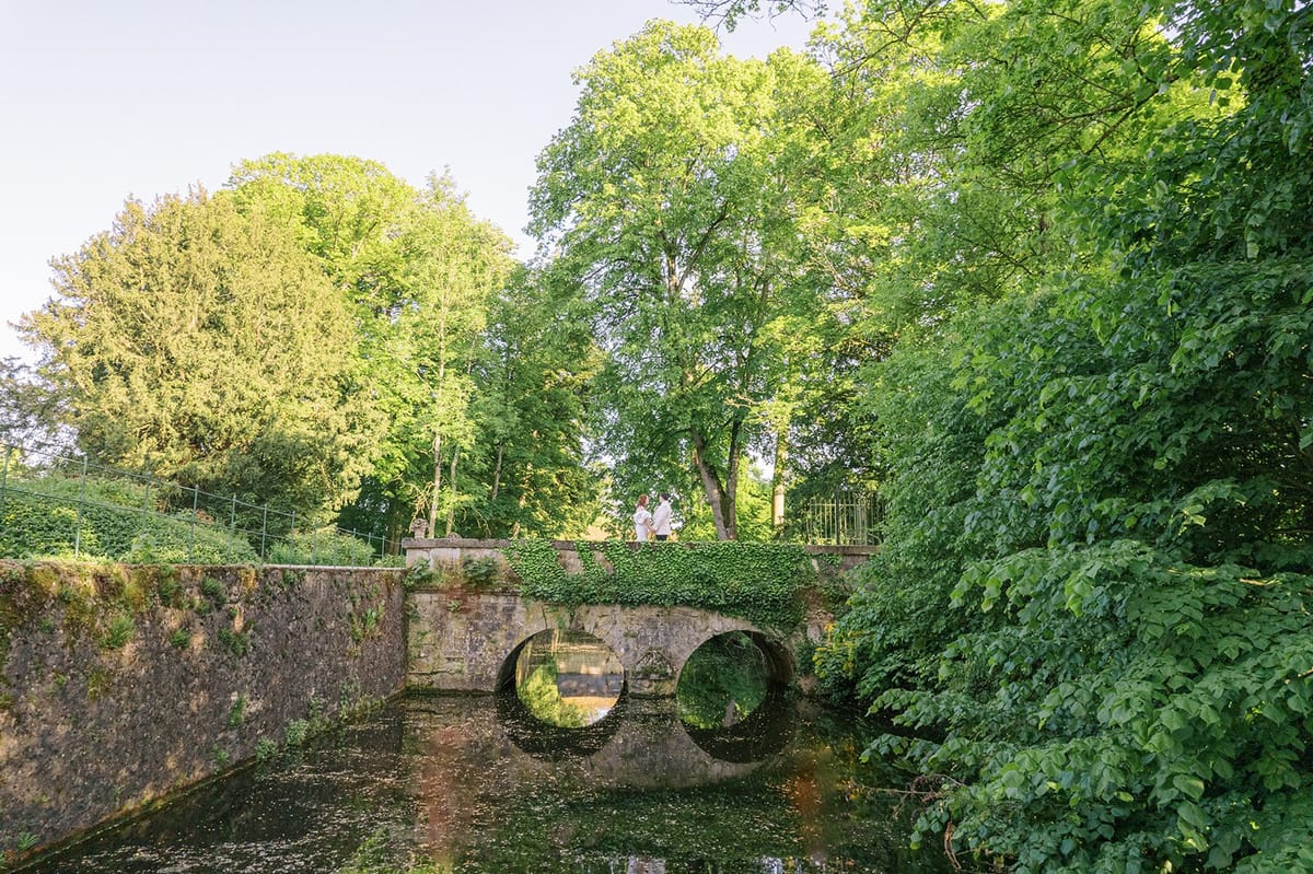 Couple posing on ivy-covered stone bridge over moat surrounded by tall trees on chateau estate grounds