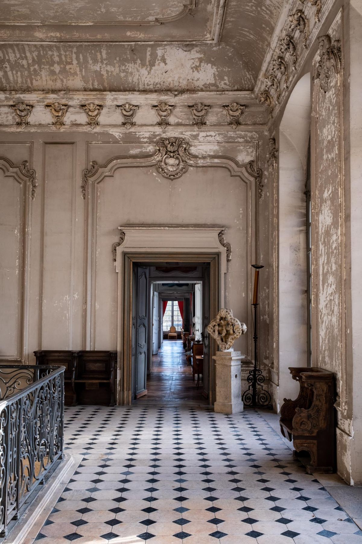 An interior architectural shot of a French château hallway with no people present. The corridor features ornate Baroque-style plasterwork moldings and carved cartouches along the upper walls and ceiling, with the plaster showing significant age-related patina and peeling. The floor is laid with a black and white diamond-pattern tile. A wrought iron balustrade runs along the left side, and antique furnishings including a carved wooden piece and a stone sculpture on a pedestal are visible along the right wall. The perspective draws the eye through a series of doorways toward a distant room hung with red curtains and a tall window. The overall palette is off-white, grey, and black, with the red curtains providing the only strong color accent at the far end. Wide shot, straight-on perspective. Potential venue feature image.