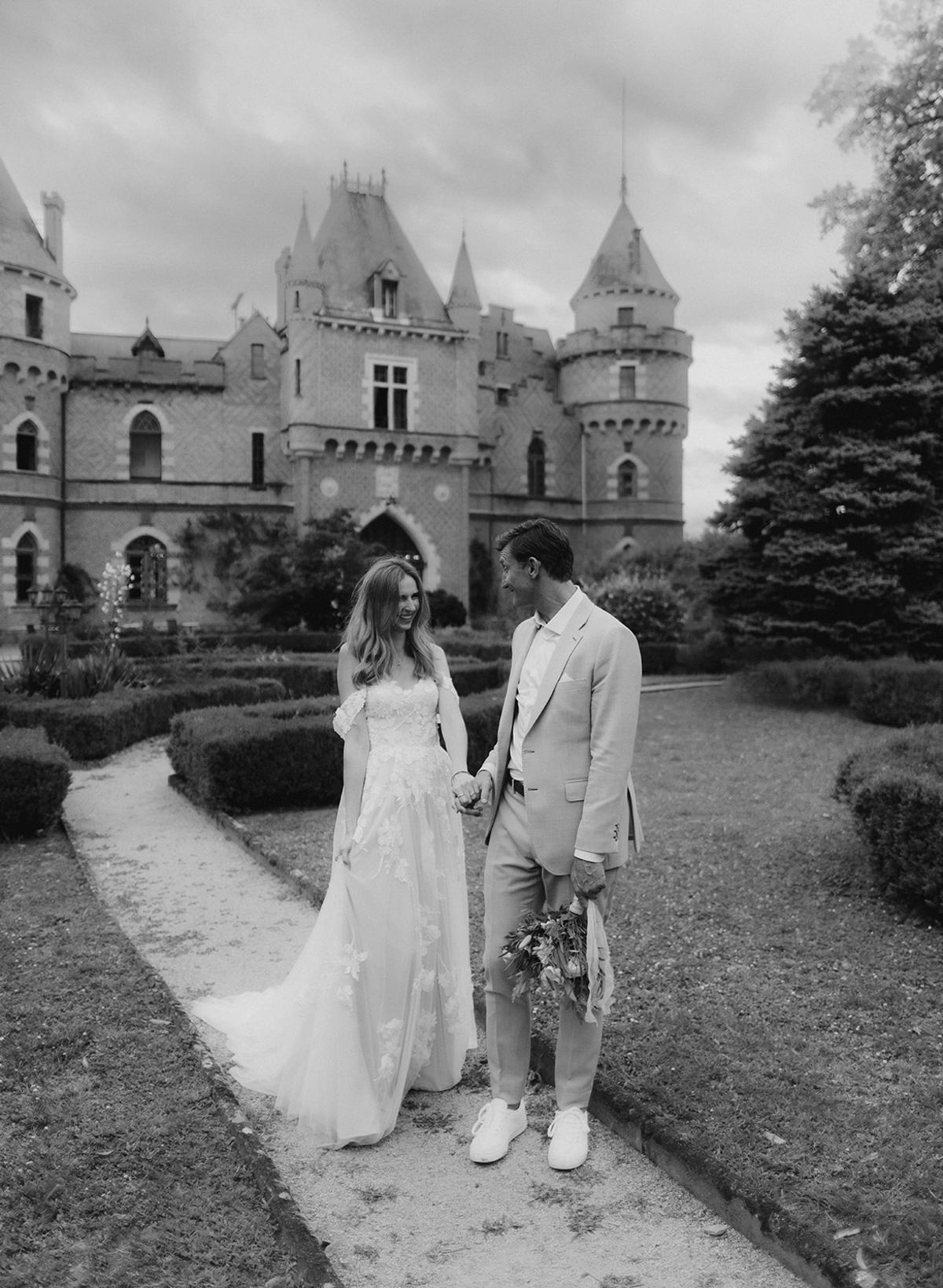Couple holds hands before Gothic-turret chateau with groom carrying trailing bouquet in B&W