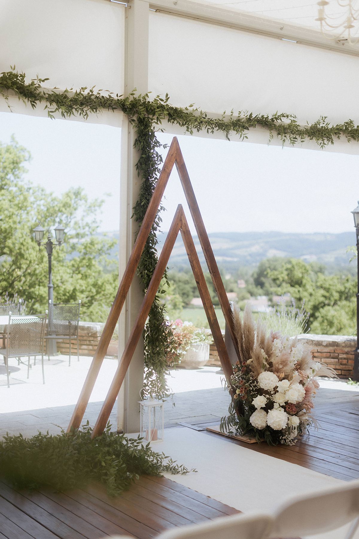 A ceremony altar setup inside a white marquee tent on a wooden deck, with an open-sided view over rolling hills and a terrace with garden furniture. The altar features two leaning triangular wooden frames forming a double A-frame arch, one dressed with trailing green foliage garlands. At the base of the arch sits a large floral arrangement combining white hydrangeas, ivory roses, blush and dusty pink blooms, pampas grass, ferns, and dried grasses in a natural, boho style. A white metal lantern sits on the deck nearby, and loose greenery is scattered along the aisle runner. Green leaf garlands also line the marquee's structural beams overhead. The overall decor palette is ivory, blush, and green with a rustic-boho styling theme. Wide shot capturing the full altar installation and surrounding venue setting.