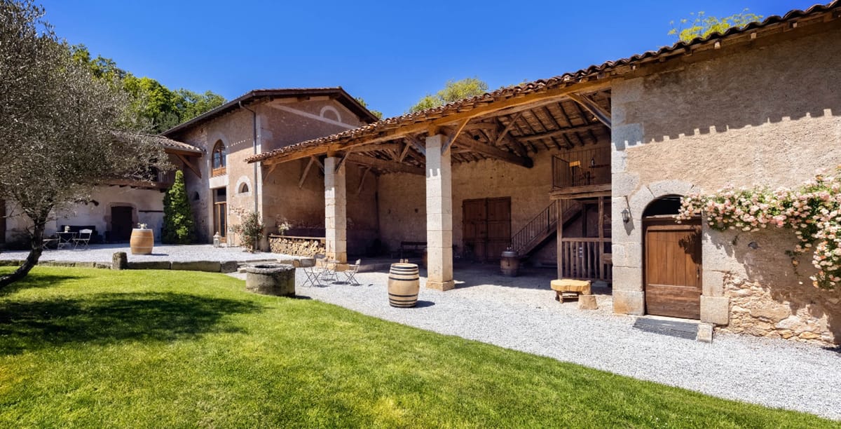 A wide exterior shot of a rustic French domaine or farmhouse property, showing a courtyard area with a gravel surface and adjacent lawn. The building features warm golden stone walls, terracotta tile roofing, and an open covered veranda structure supported by stone columns with exposed wooden beam rafters. Decorative details include a climbing rose bush with soft pink blooms framing a heavy wooden arched door on the right facade, and a wrought iron staircase leading to a mezzanine level. Wine barrels are used as decorative props throughout the courtyard, and a stone well sits centrally near small metal bistro chairs and a table. A terracotta-colored large barrel and a wood stump side table add to the rustic styling. No people are visible in the image. Potential venue feature image.