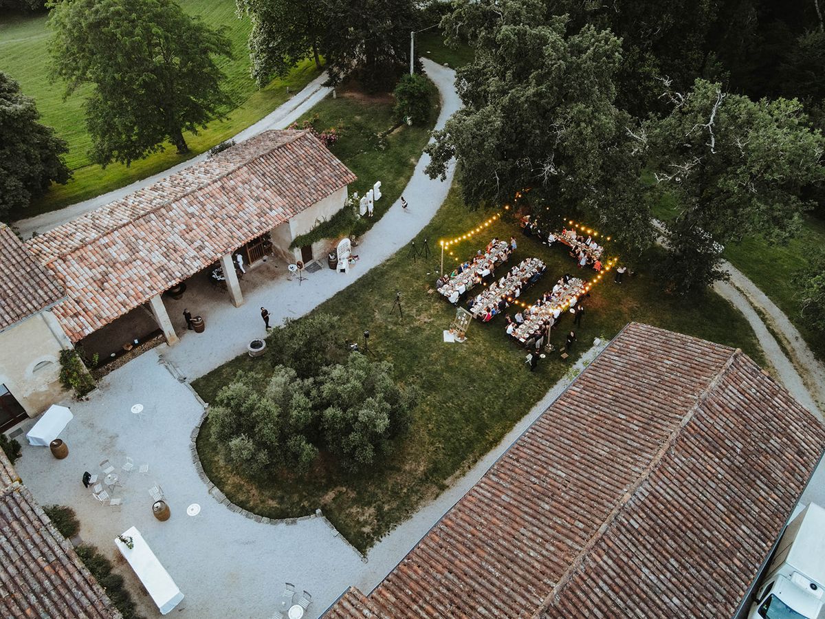 Aerial dusk reception three parallel tables on estate lawn with festoon lights stone outbuildings and gravel courtyard