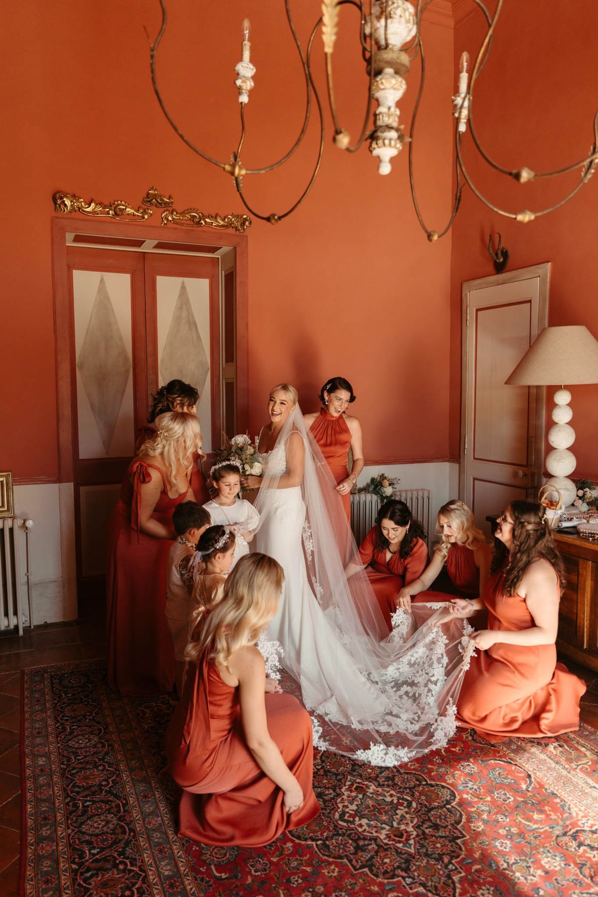 A getting-ready scene captured indoors in a grand room with deep terracotta-red painted walls, ornate gold-leaf door moldings, an iron chandelier, and a patterned red oriental rug on a stone floor. The bride stands at the center in a fitted white lace gown with a cathedral-length veil edged in appliqué lace detailing, holding a small bouquet of soft white and blush flowers, laughing as she is surrounded by her bridal party. Six bridesmaids wear burnt orange or terracotta floor-length satin dresses in varying styles including halter and sleeveless cuts, with several kneeling or sitting on the floor to arrange and spread the veil train. Two young flower girls in white dresses with floral hair accessories stand near the bride. The composition is a wide shot taken from a slightly elevated angle, capturing the full scene including the room's architectural details and decor.