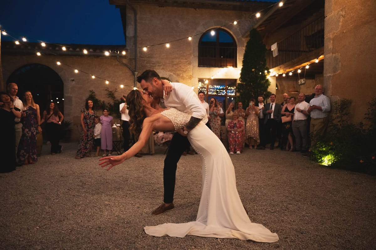 Bride and groom first dance dip under string lights in rustic stone courtyard venue at night with guests watching