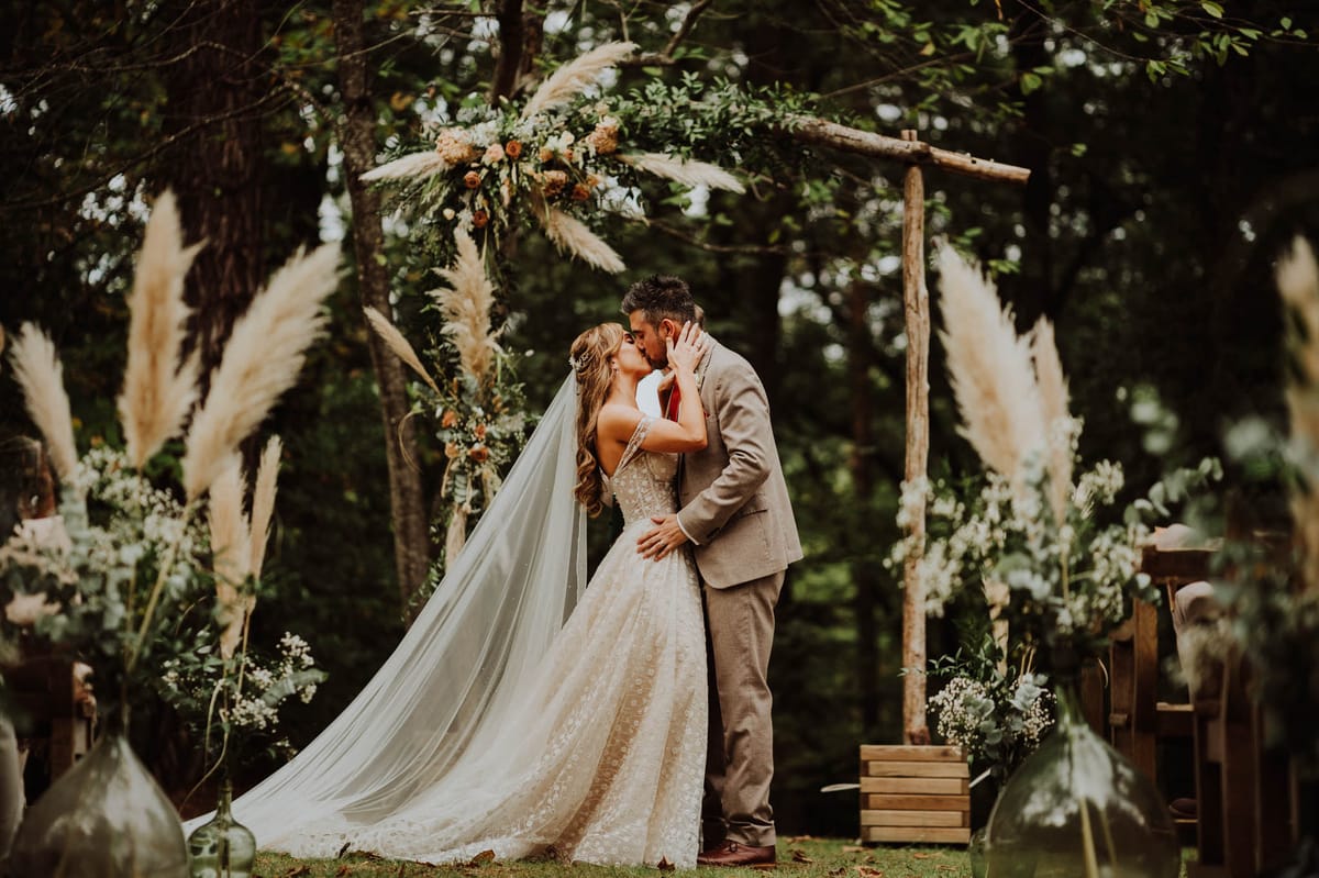 The bride and groom share their first kiss during an outdoor woodland ceremony, framed by a rustic wooden arch made from natural branches. The bride wears a lace ball gown with a low back and a long cathedral-length veil, while the groom is dressed in a taupe linen suit. The arch is decorated with pampas grass, greenery, burnt orange and terracotta ranunculus, and white florals, carried through to floor-level arrangements along the aisle, which also feature glass carboys, wooden crates, and baby's breath clusters. The overall styling is boho-rustic with a warm, earthy color palette of terracotta, ivory, and sage green. This is a wide portrait shot with a shallow depth of field, with foreground pampas grass framing the couple.