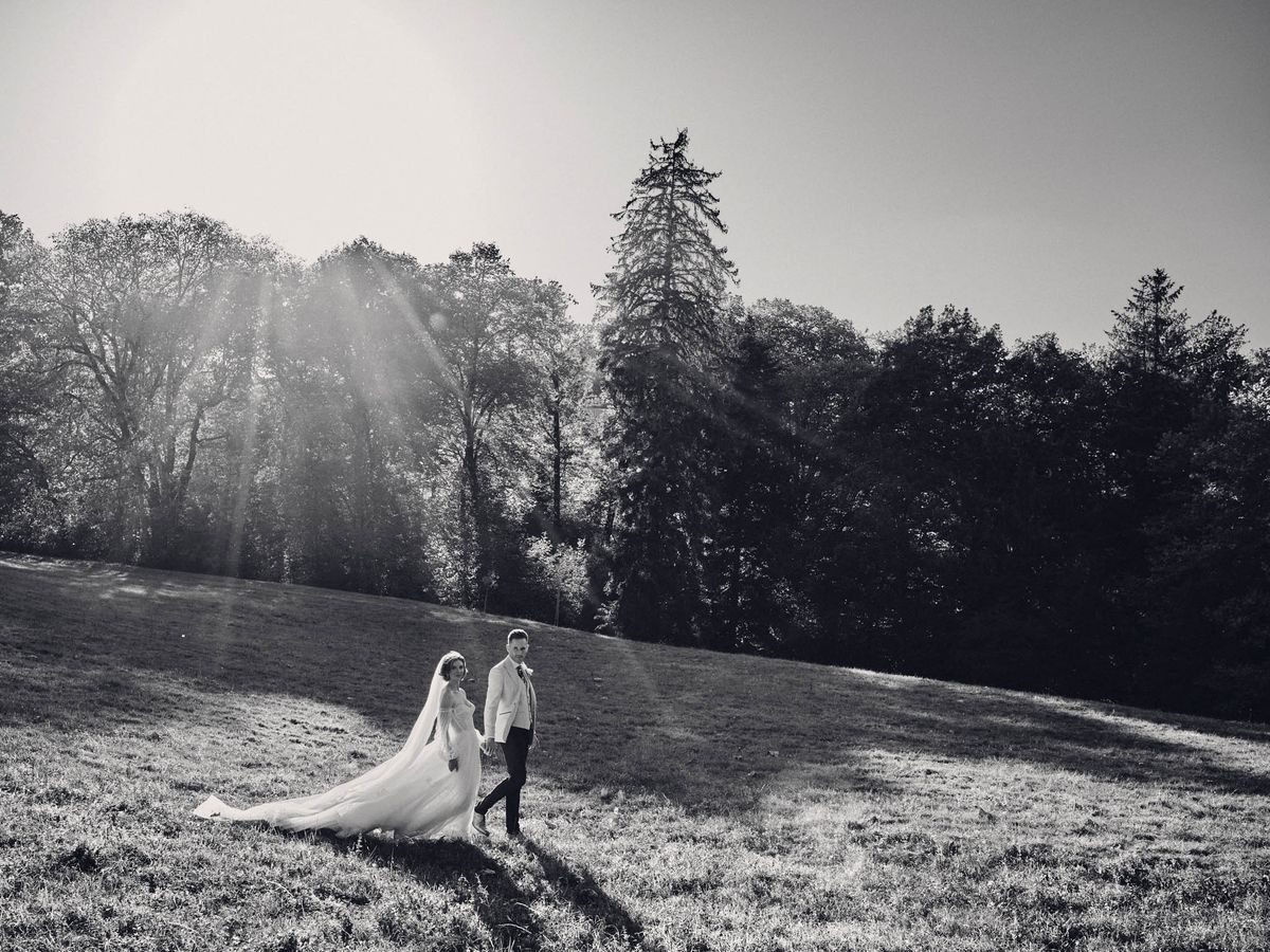 This is a black-and-white couple portrait taken outdoors in a wide open parkland setting. The bride and groom are walking hand in hand across a sloped grassy field, positioned small within the frame to emphasize the expansive landscape around them. The bride wears a full-length gown with a very long cathedral-length veil that trails behind her across the grass, while the groom is dressed in a light-toned suit with dark trousers. Strong sun flare bursts through the trees in the upper left of the frame, creating high contrast between the bright sky and the deep shadow tones of the tree line in the background. The composition is a wide environmental shot with the couple anchored in the lower-center third, conveying scale and movement.