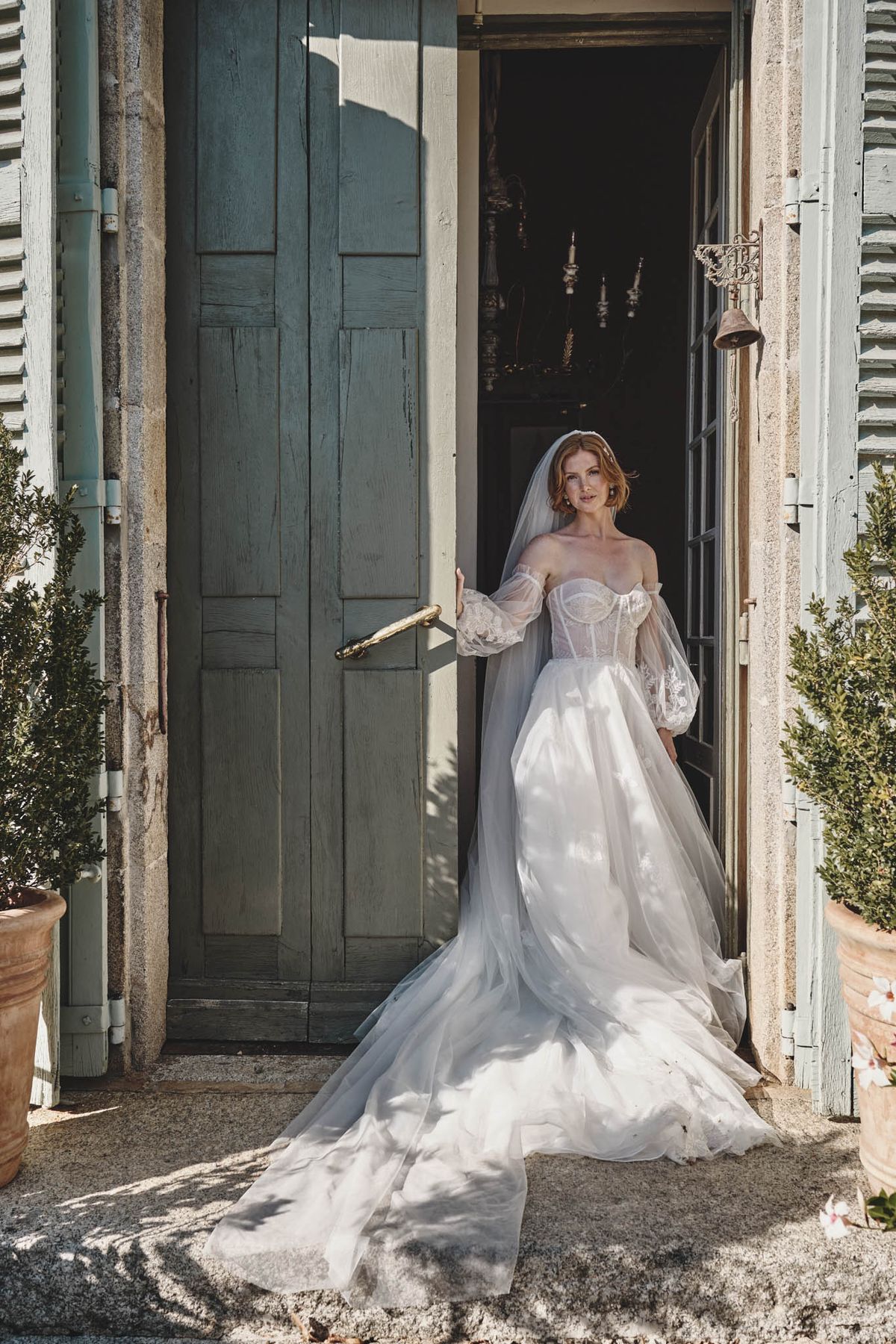 A bridal portrait of a single bride standing in the open doorway of what appears to be a French château or manor house, shot as a full-length portrait in natural sunlight. The bride wears an off-the-shoulder ball gown with a sheer lace corset bodice, voluminous puff sleeves in organza or tulle, and a full layered skirt with an extended train spread across the stone threshold. A cathedral-length veil is worn over her short auburn hair, and she holds one of the aged sage-green painted wooden doors open. In the darkened interior behind her, a candelabra chandelier is faintly visible. Two boxwood topiaries in terracotta pots flank the entrance, and the overall styling has a romantic, vintage-influenced aesthetic. Potential venue feature image.