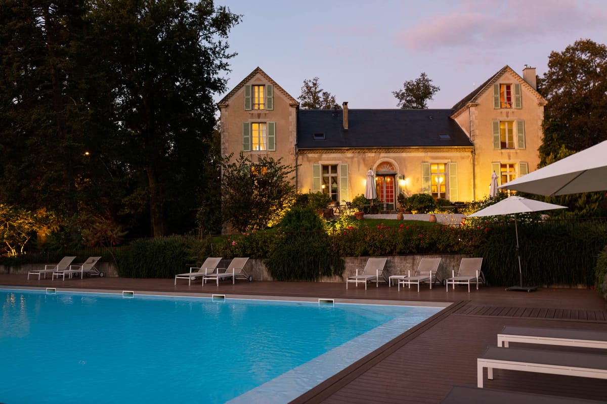 Wide-angle exterior shot of a French stone manor house photographed at dusk, with warm interior lighting glowing through windows fitted with sage green shutters and a red arched front door. The two-storey limestone building with a dark slate roof is set back behind a landscaped garden with warm uplighting illuminating the shrubbery and flowering plants in the foreground. In the foreground, a large rectangular swimming pool with built-in blue underwater lighting is bordered by a dark wood deck lined with white sun loungers and white patio umbrellas. No people are visible in the image. Potential venue feature image.