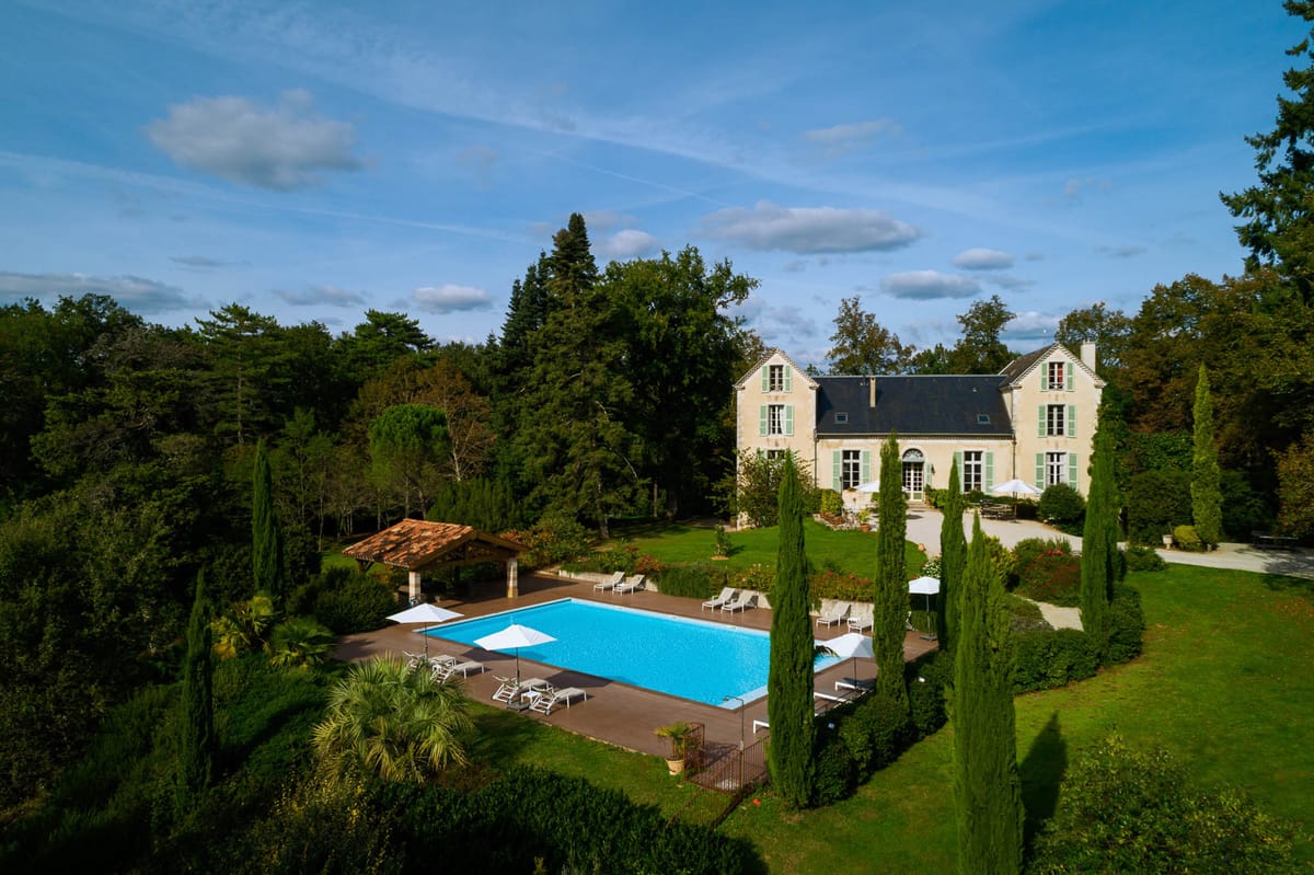 An aerial wide shot of a French country manor featuring a cream-rendered façade with sage green shutters, a dark slate mansard roof, and multiple large windows. The grounds include a large rectangular outdoor swimming pool with a terracotta-roofed poolside pavilion, white sun loungers, and white market umbrellas arranged around a dark-toned pool deck. A gravel driveway curves past the front of the building, flanked by tall columnar cypress trees and manicured lawn areas with rose plantings. No people are visible in the image. Potential venue feature image.