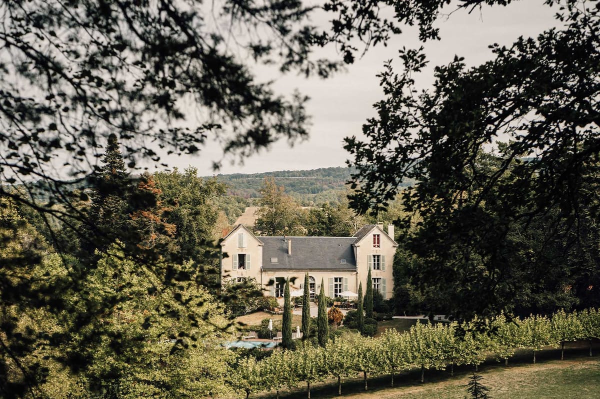 Wide exterior shot of a French manor house viewed from a distance through a frame of tree branches, with no people visible. The two-story cream-rendered building features a dark slate roof, pale green shutters, tall arched windows, and chimneys consistent with classic French provincial architecture. A swimming pool, white market umbrellas, outdoor seating, and a row of tall cypress trees are visible in the foreground grounds, while wooded hills extend across the background. The composition is taken from an elevated vantage point, with overhanging branches creating a natural frame around the property. Potential venue feature image.